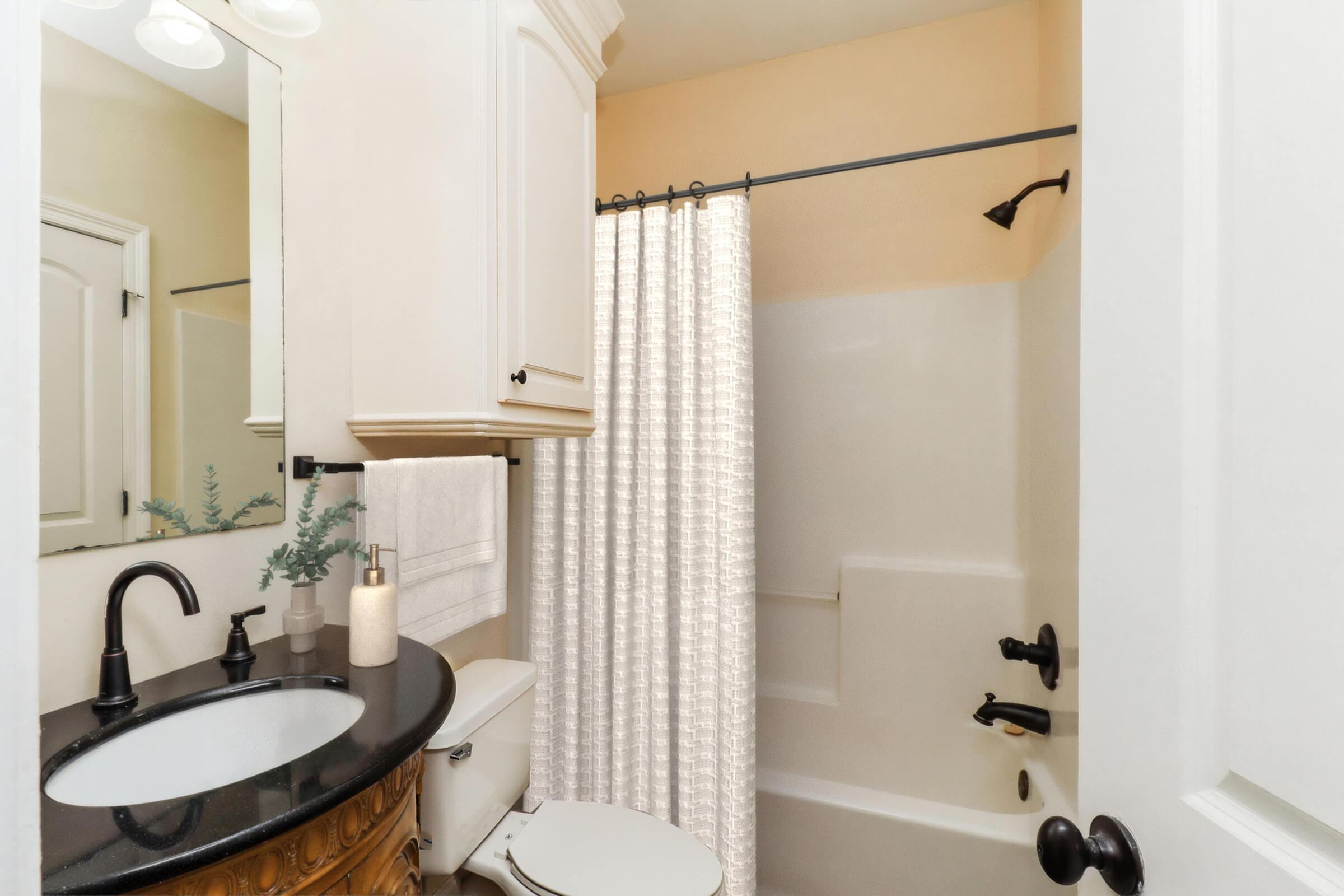 A small bathroom featuring a white bathtub with a shower curtain, a dark granite countertop sink with a faucet, and a wooden vanity. Light-colored walls and a towel rack are visible. The overall design is clean and modern, with decorative elements like a plant on the countertop.
