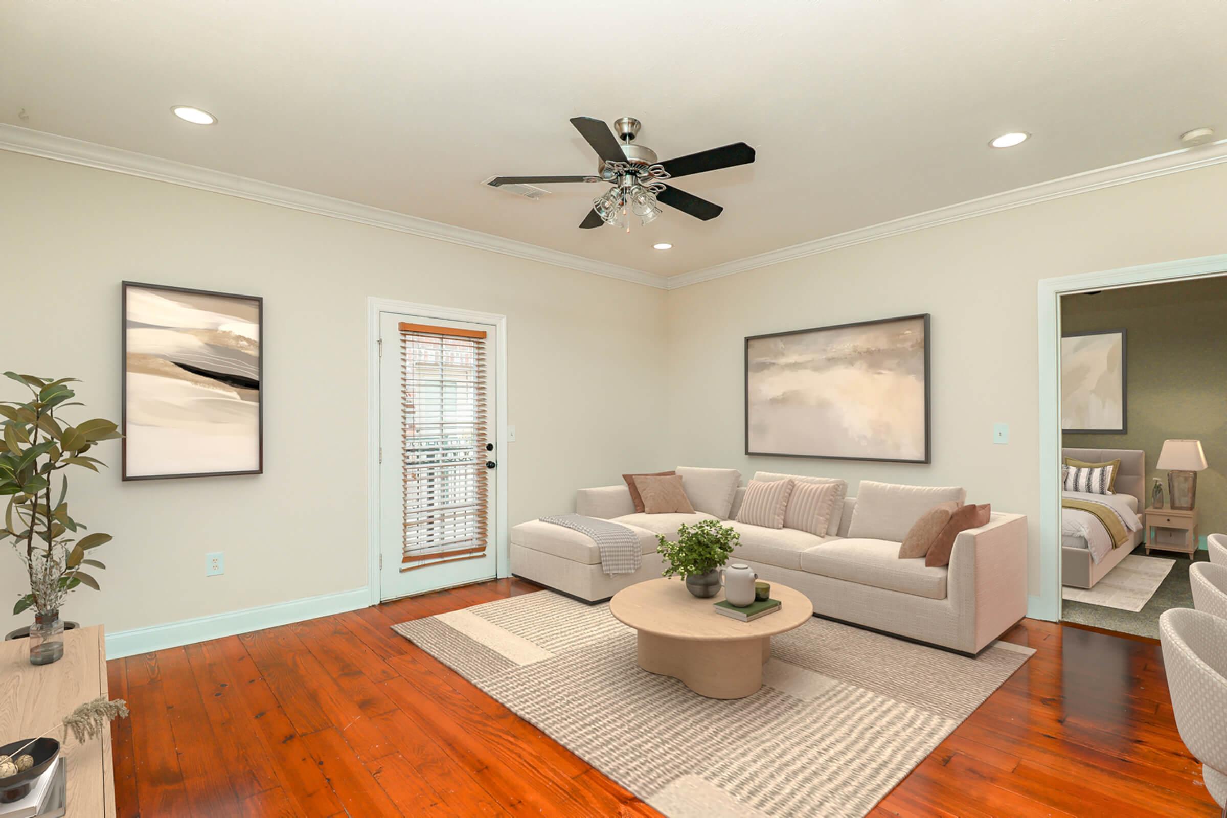 A modern living room featuring a light-colored sofa with cushions, a round coffee table, and decorative plants. The walls are painted a soft color with two abstract paintings, and there’s a door leading outside. The space has hardwood flooring and a ceiling fan, creating a cozy and inviting atmosphere.