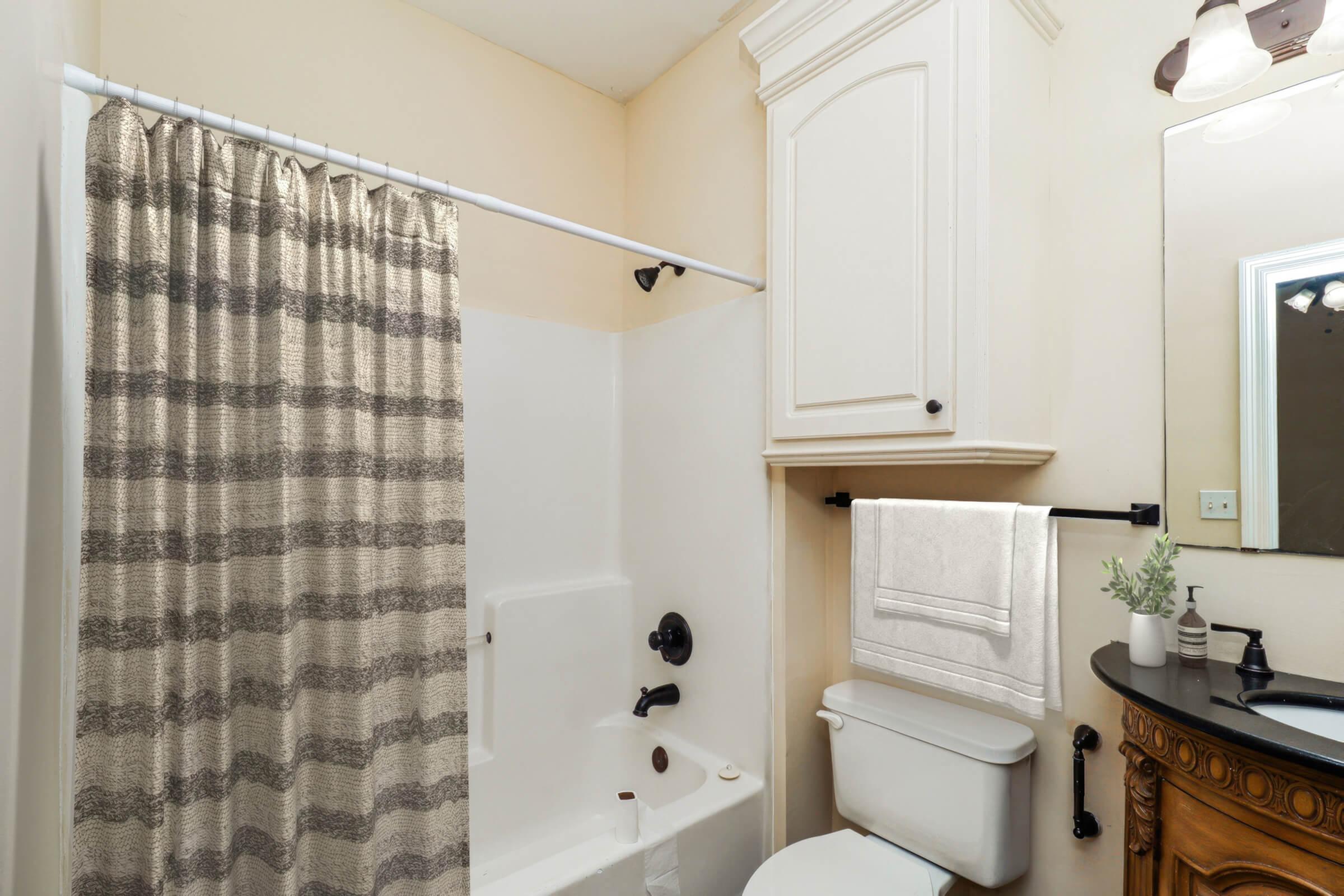 A clean, well-lit bathroom featuring a shower with a striped curtain, a white bathtub, a toilet with a wooden cabinet above it, a towel hanging on a rack, and a round mirror above a dark countertop. The walls are painted a light color, creating a spacious feel.