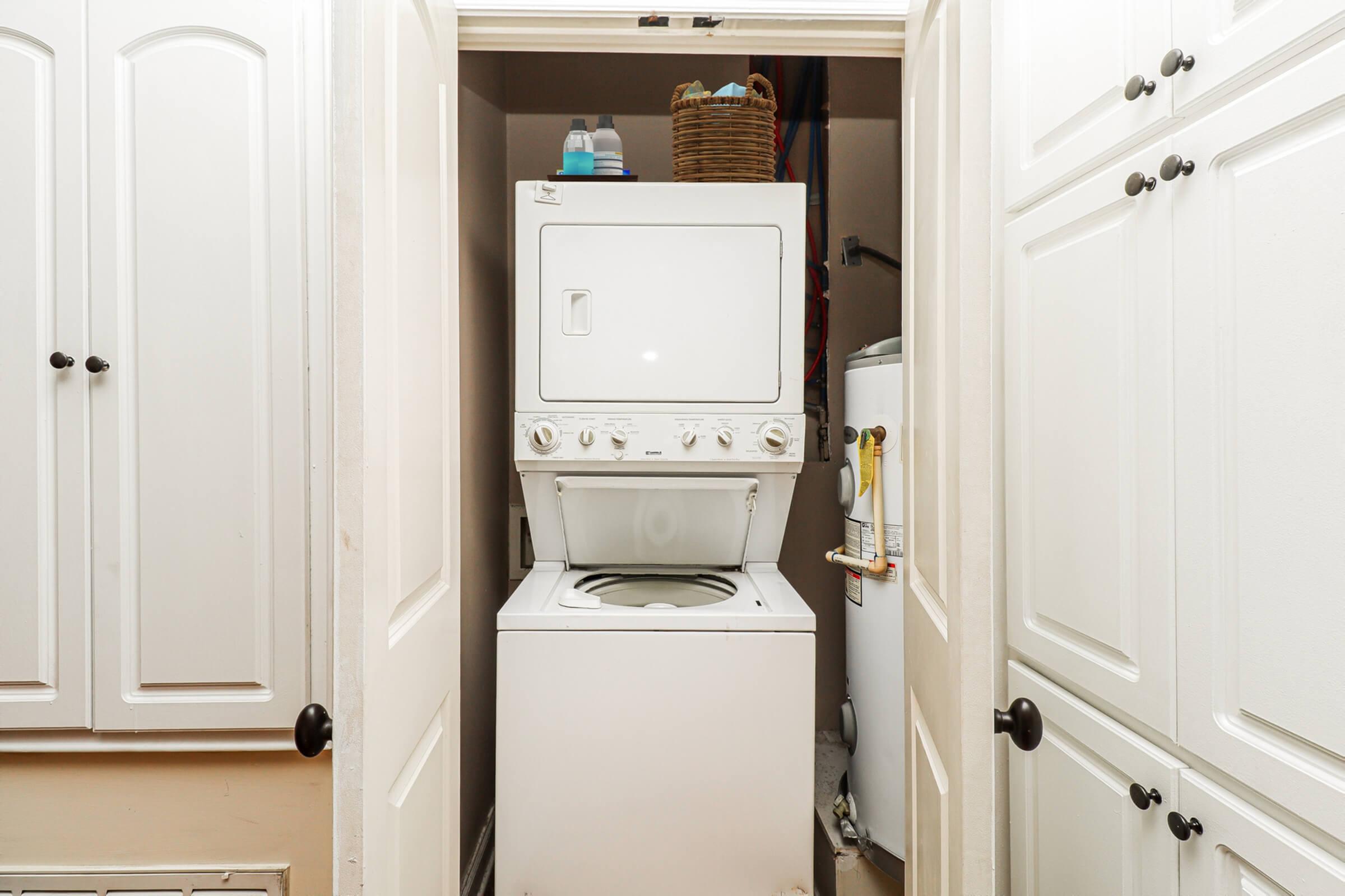 A stacked washer and dryer are positioned between two open white doors, leading into a laundry area. A woven basket sits on top of the dryer, and a water heater is visible in the corner, contributing to a compact and functional laundry space.