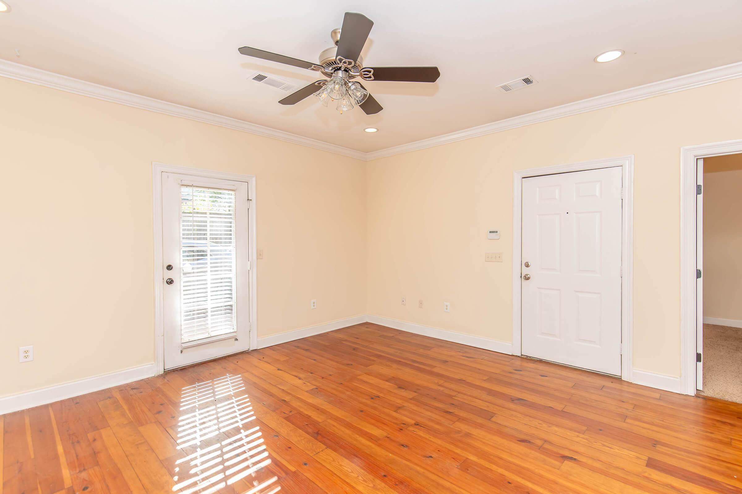 A spacious, empty room with light hardwood flooring, cream-colored walls, and a ceiling fan with lights. There are two doors: one leading to an outdoor area with blinds and one leading to another room. Natural light is streaming in, creating a warm and inviting atmosphere.