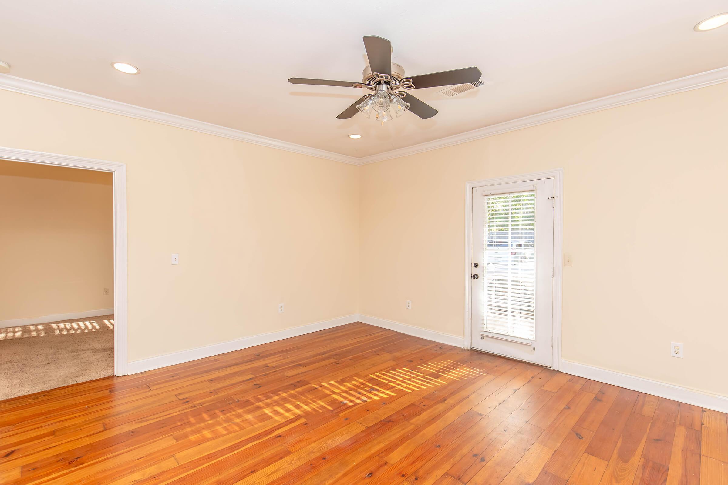 A spacious, well-lit room with a ceiling fan. The walls are painted a soft yellow, and the floor features polished wooden planks. A single door leads to an outdoor area, and there's an entrance to another room on the left. The overall ambiance is inviting and open.