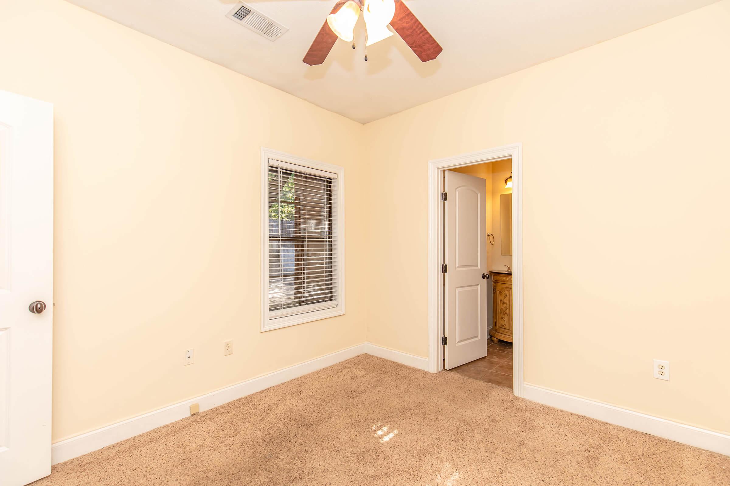 A cozy room with beige walls and carpet flooring. A ceiling fan hangs from the ceiling, and a window with blinds allows natural light. A doorway leads to a bathroom. The overall atmosphere is warm and inviting, suitable for a bedroom or office space.