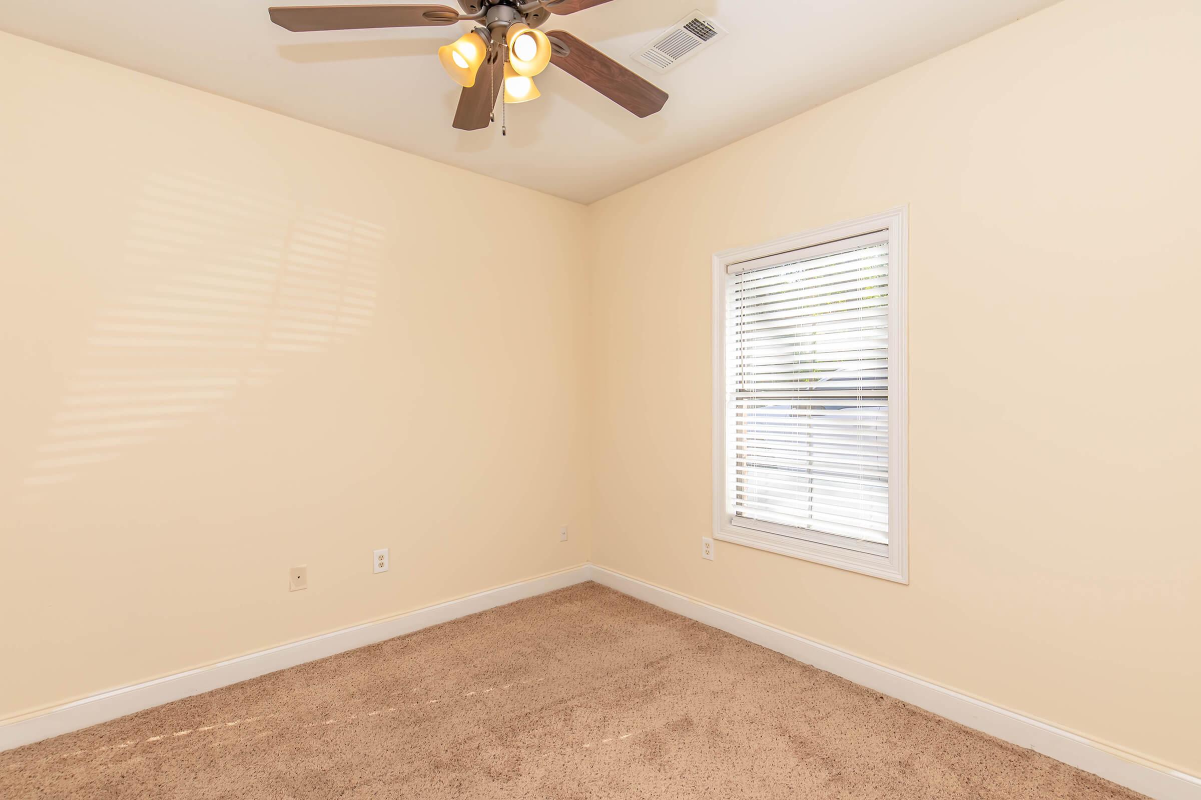 Empty room with light yellow walls, a ceiling fan with four lights, and a window with blinds letting in natural light. The floor is covered with beige carpet, and there are no furnishings or decorations visible.