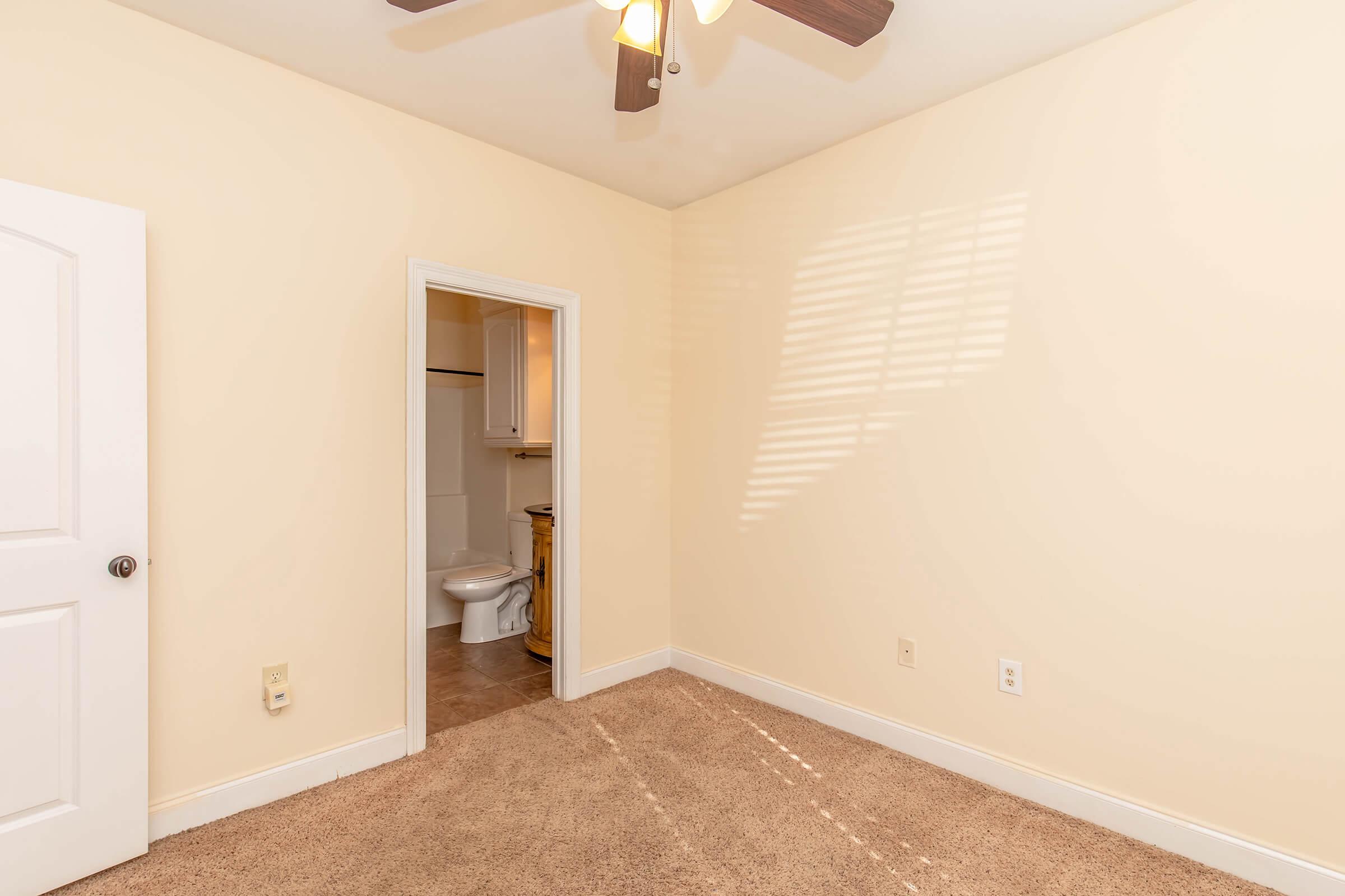 A small, empty bedroom featuring light-colored walls, carpeted floor, and a ceiling fan. There is a door leading to a bathroom area visible through an open doorway. Natural light casts shadows across the room from a nearby window.