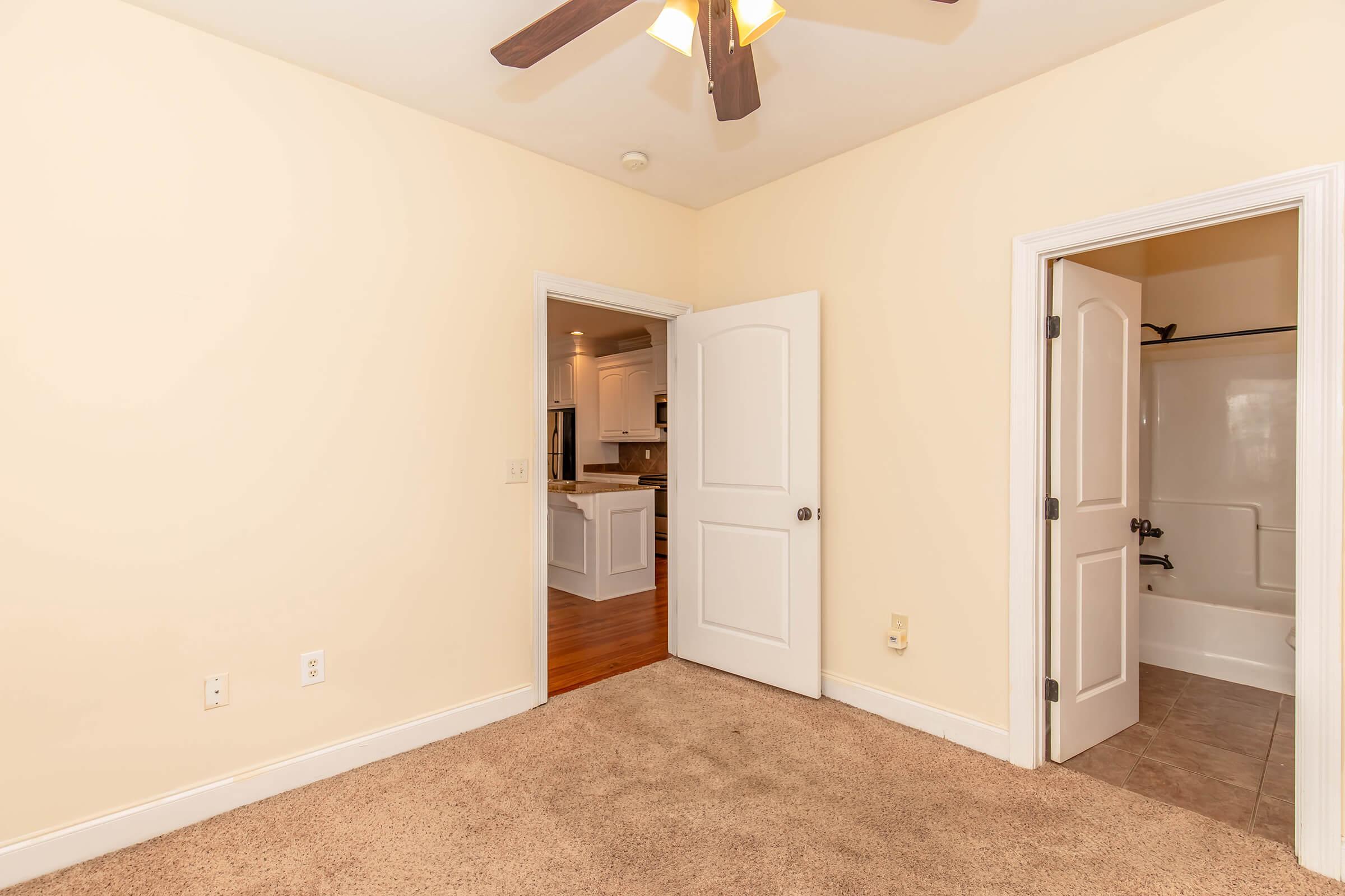 A small, well-lit room with beige walls and a ceiling fan. The room features a plush carpet and two open doorways leading to adjacent spaces. One doorway opens to a bathroom with a tiled floor, while the other leads to a kitchen area. Natural light enhances the inviting atmosphere.