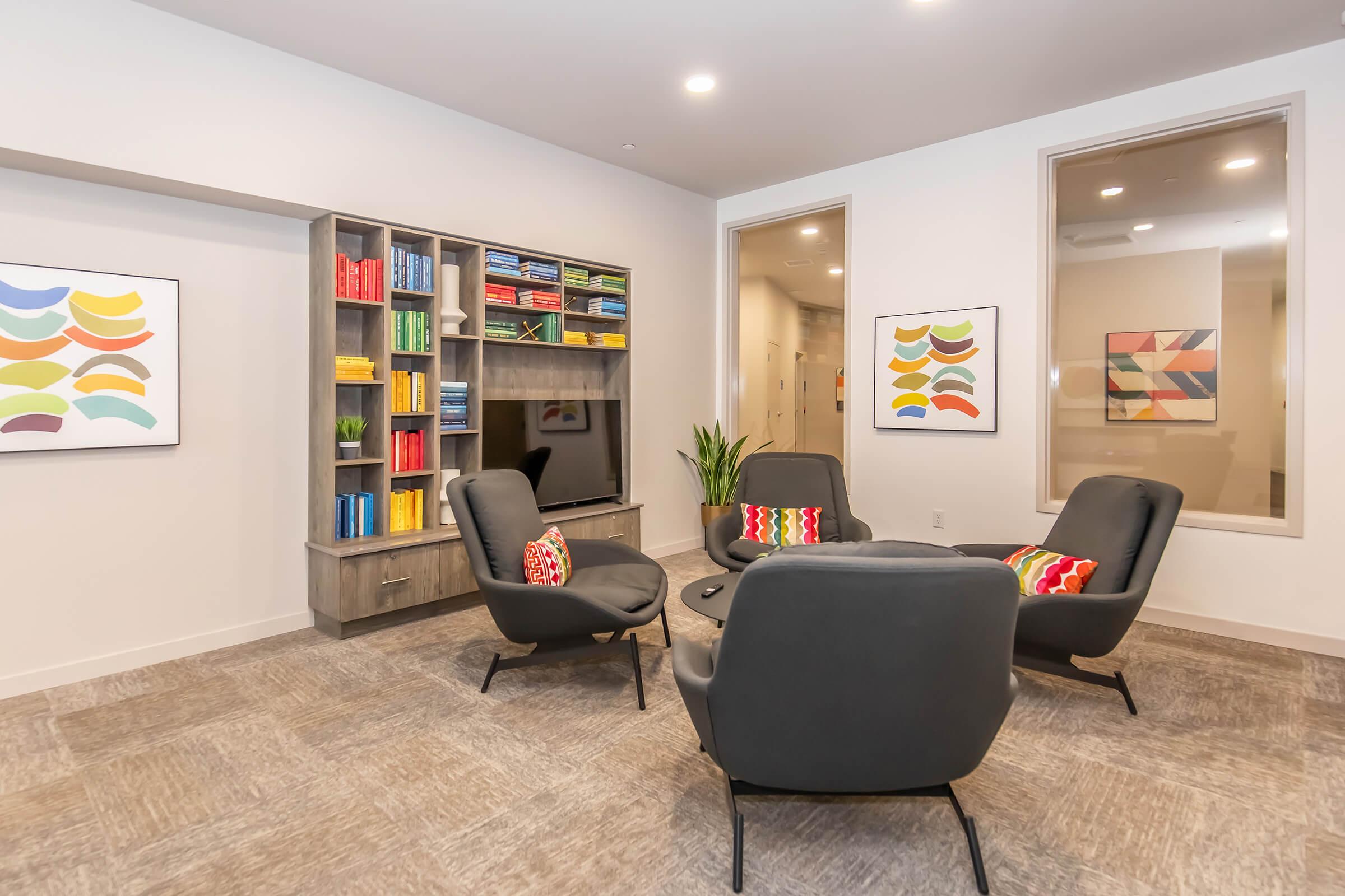 A cozy lounge area featuring four modern chairs arranged around a circular table. A bookshelf filled with colorful books is against one wall, accompanied by two abstract art pieces. The space is well-lit with large windows, creating a welcoming atmosphere for relaxation or conversation.