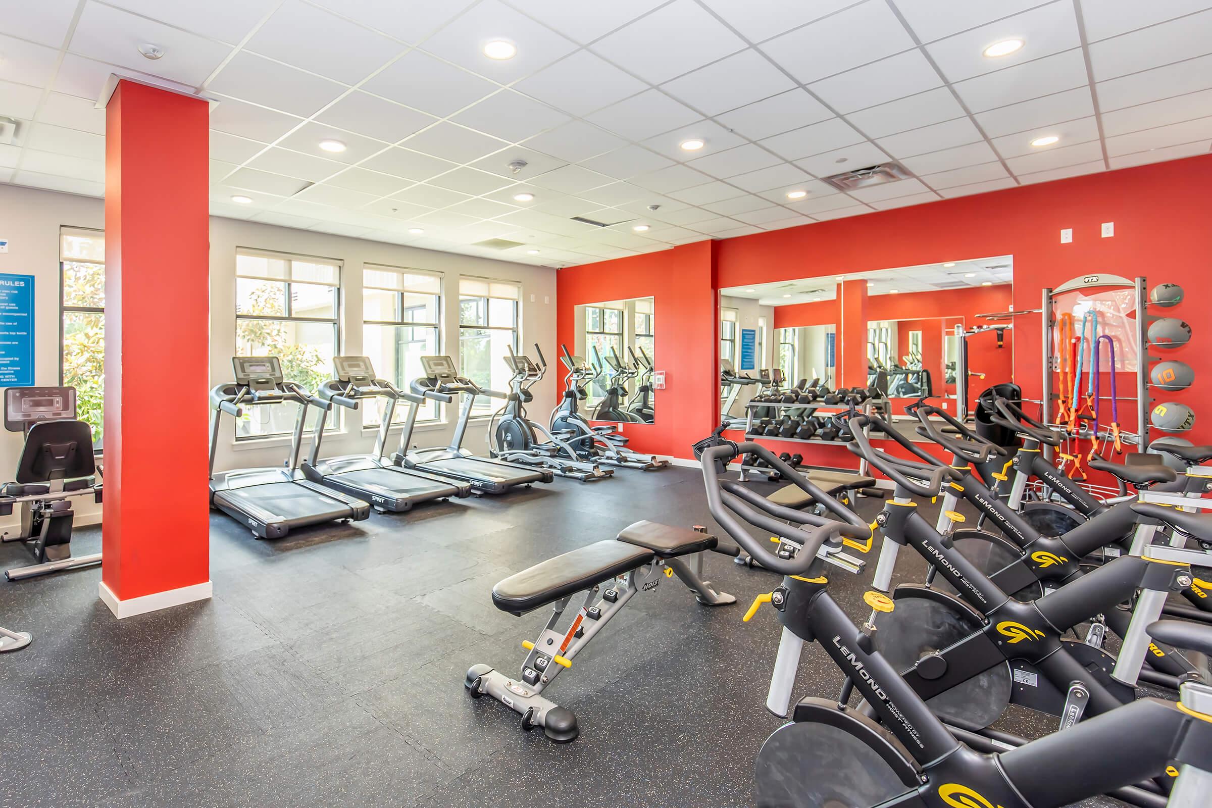 A modern gym interior featuring multiple treadmills and weight training equipment. The room has red accent walls, large windows allowing natural light, and mirrors reflecting the workout area. Fitness accessories and exercise mats are visible, creating a vibrant and inviting workout environment.