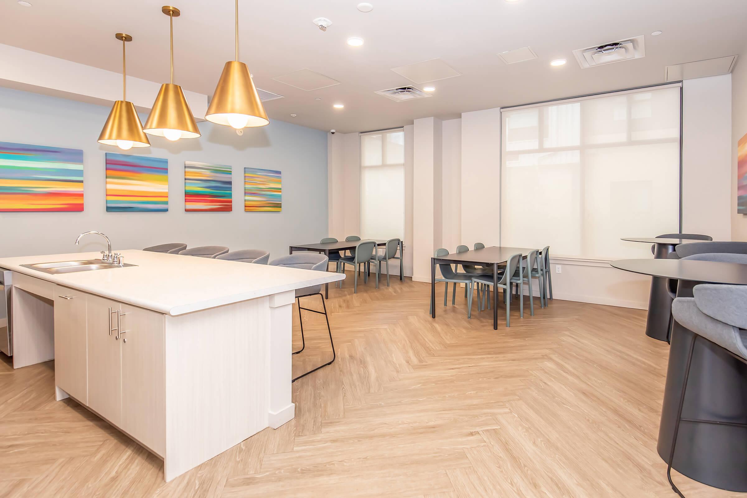 A modern, spacious kitchen and dining area featuring a large island with a sink, surrounded by gray chairs. The walls are adorned with colorful abstract paintings, and large windows allow natural light to fill the room. There are several dining tables arranged, with sleek bar stools nearby.