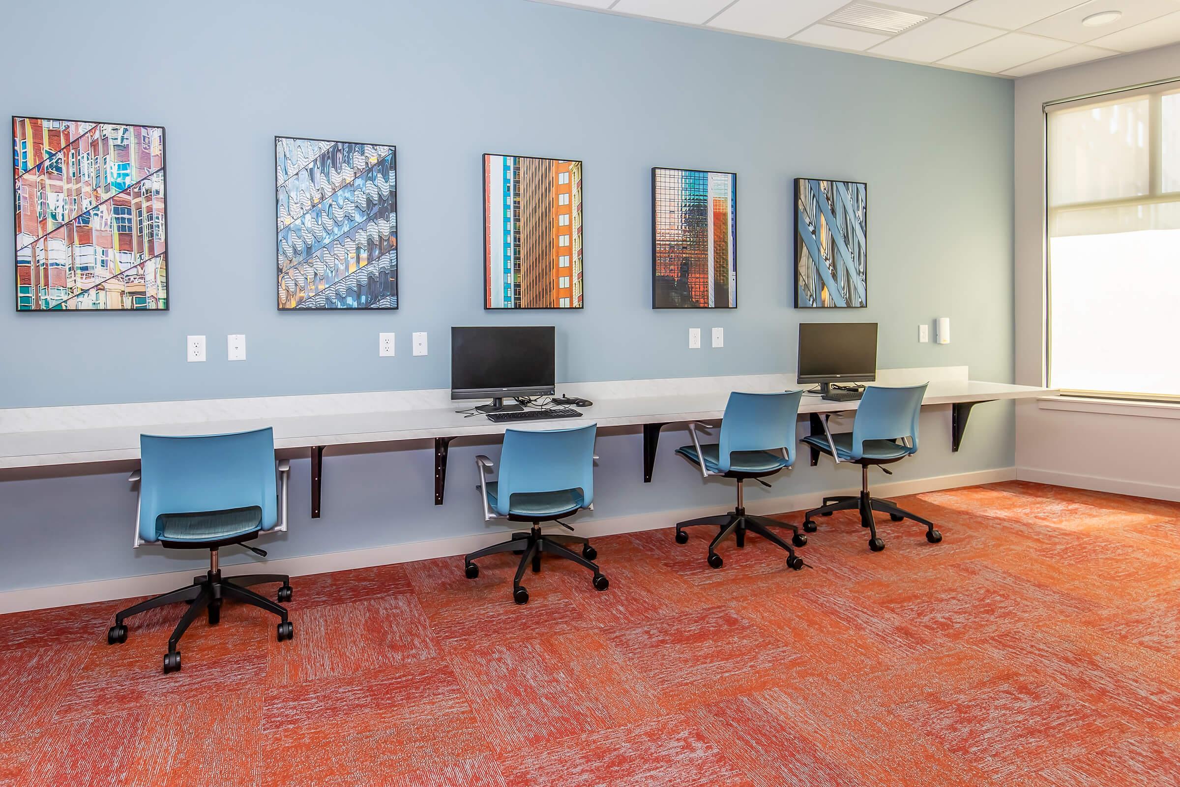A modern workspace featuring a long white desk with four computer stations and rolling chairs. The walls are adorned with vibrant framed artwork, and the room has a bright, inviting atmosphere with orange carpet and large windows allowing natural light.
