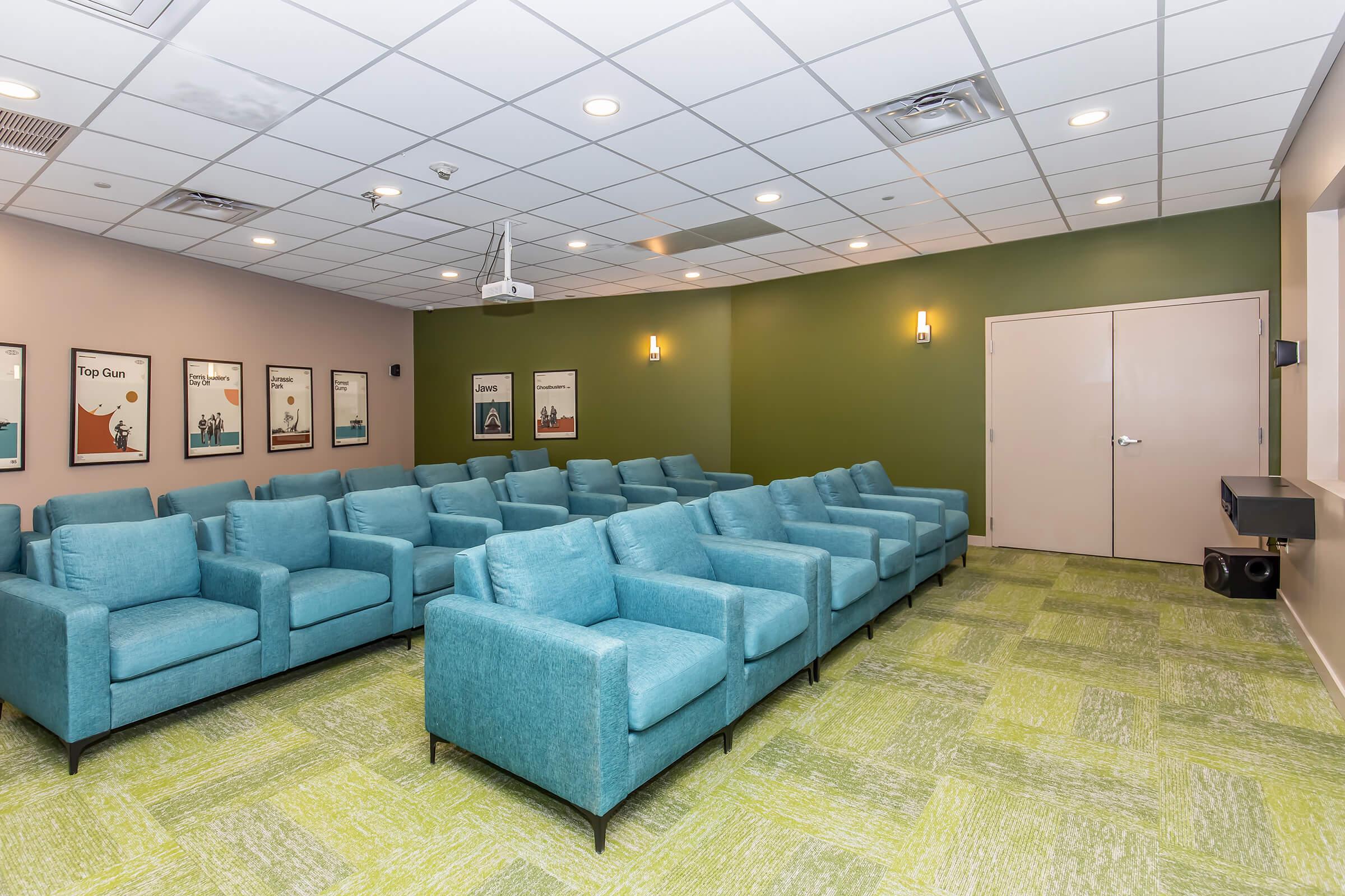A comfortable movie theater room featuring rows of teal upholstered chairs, a green accent wall, and framed movie posters on the walls. The ceiling is equipped with lighting and a projector setup, creating an inviting atmosphere for movie screenings or presentations.