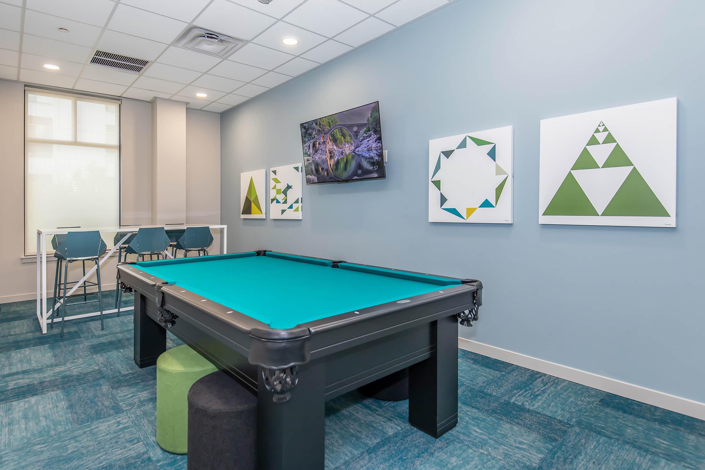 A game room featuring a billiard table with a teal felt surface. On the walls are modern geometric art pieces, and a flat-screen TV mounted above. There are four stylish stools arranged around a glass-topped table in the corner, enhancing the contemporary decor of the space with blue carpet.