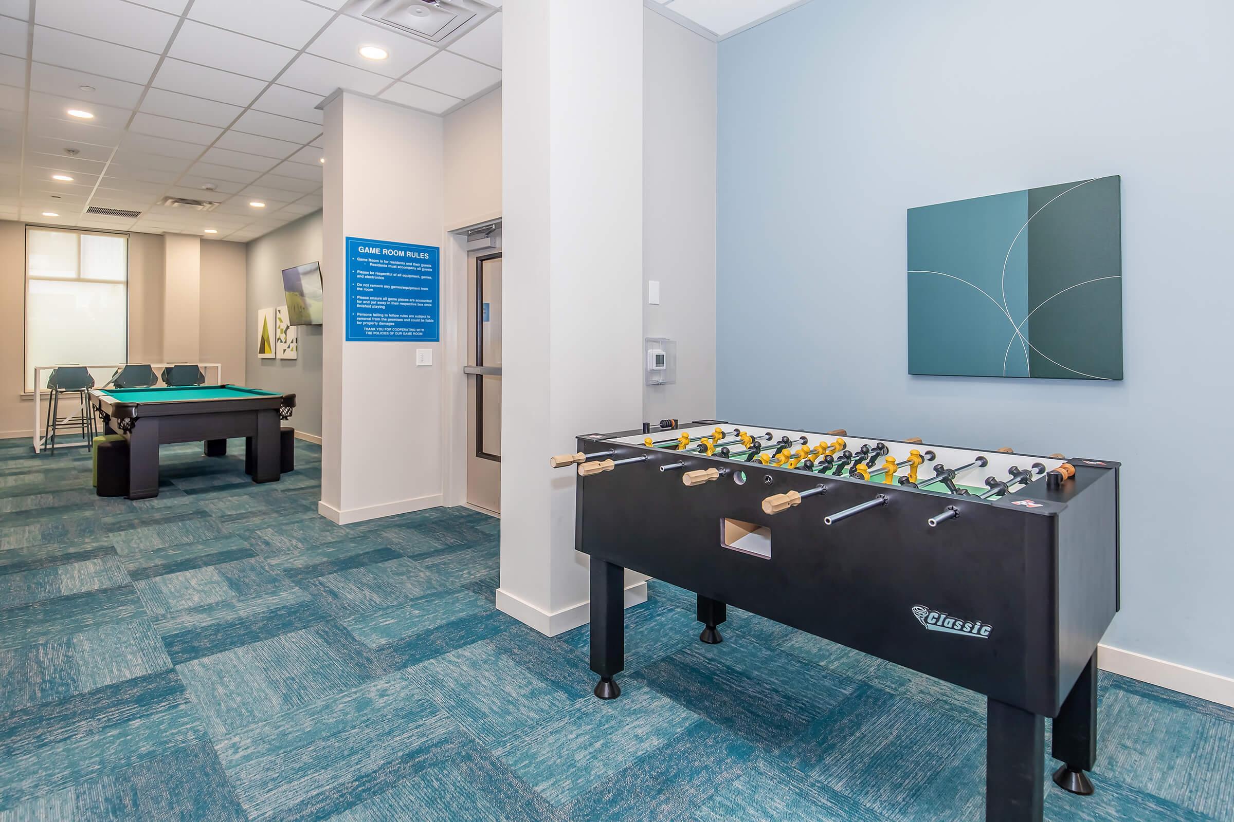 A modern game room featuring a foosball table and a pool table in the background. The room has light blue walls and carpeted flooring, with a minimalist artwork on one wall and informational signage. It's designed for recreational activities and socializing.