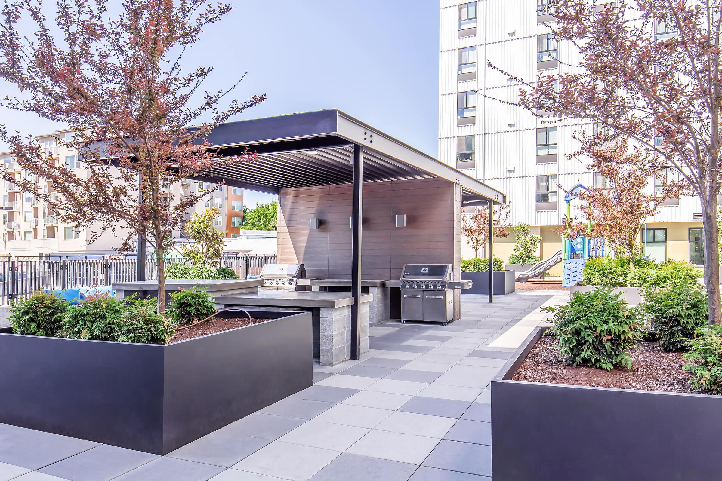 Outdoor kitchen area featuring a covered grill station with stainless steel appliances. Surrounding green landscaping includes small trees and shrubs, set against a modern urban backdrop with apartment buildings. The space is designed for social gatherings and relaxation.