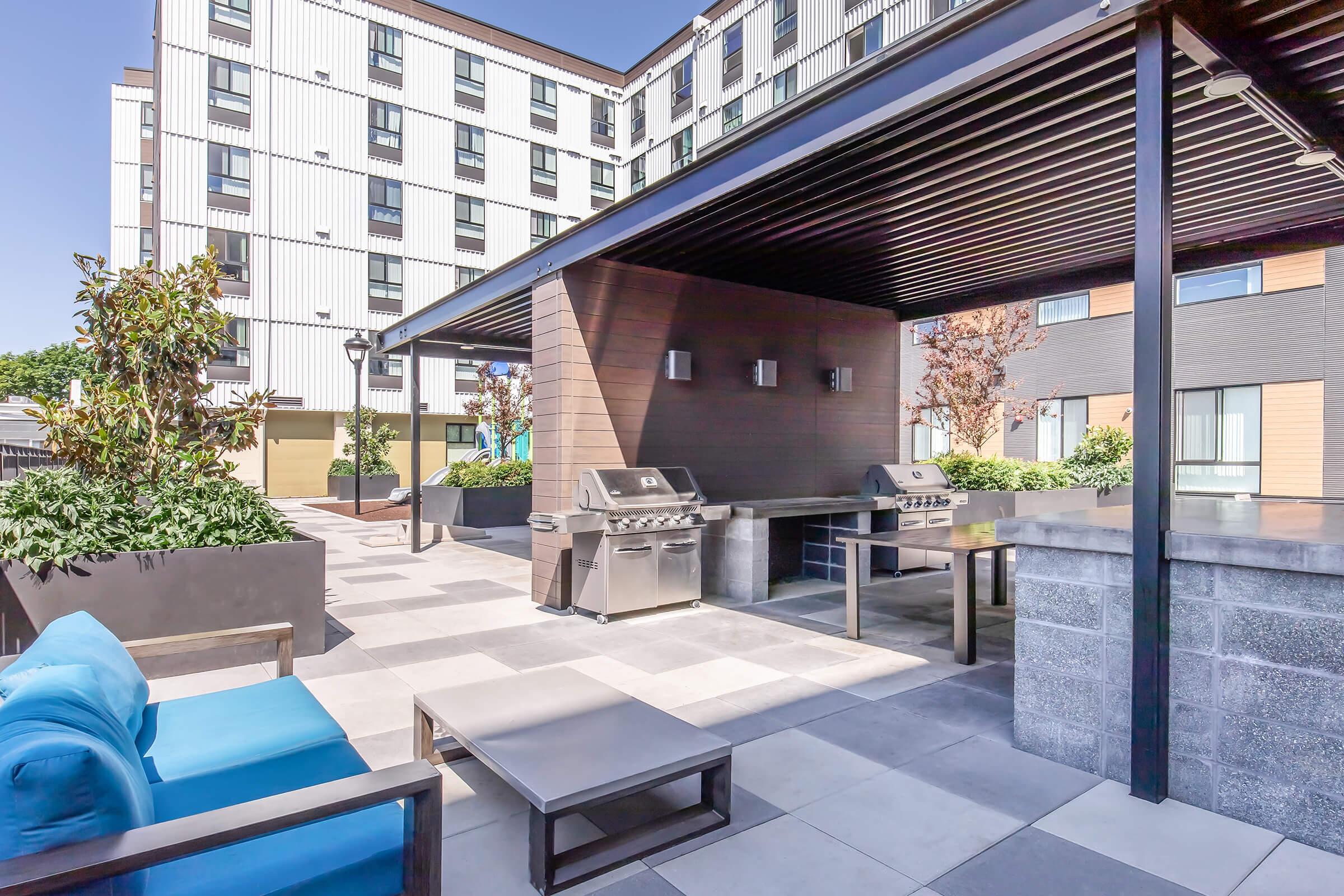 Outdoor patio area featuring a barbecue grill station, modern seating with a blue couch, and tables. The backdrop includes a multi-story building with contemporary architecture, surrounded by landscaped plants and trees under a clear blue sky.