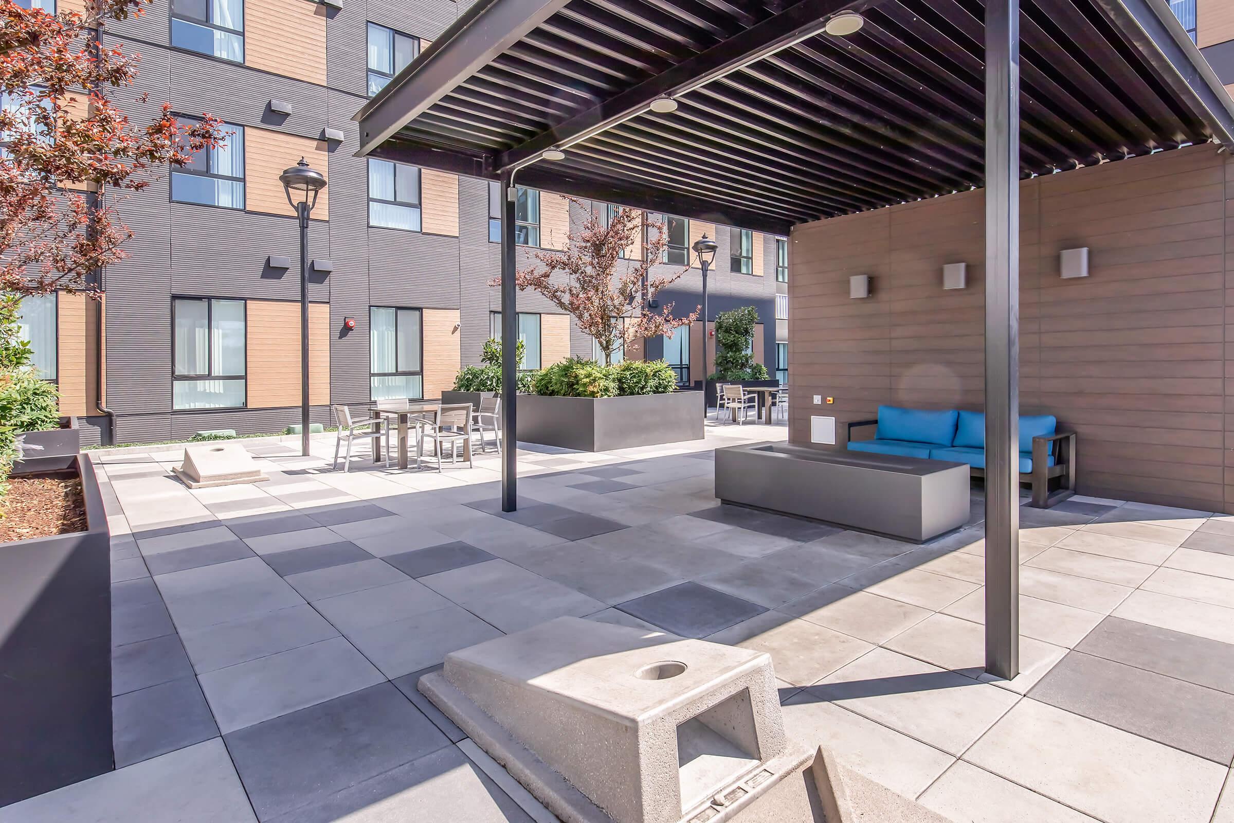 A modern outdoor lounge area featuring a shaded seating space with blue cushions, surrounded by planters and tables. The setting includes stone pavers and is adjacent to a contemporary building with a mix of dark and light siding. Trees and landscaping add a touch of greenery to the environment.
