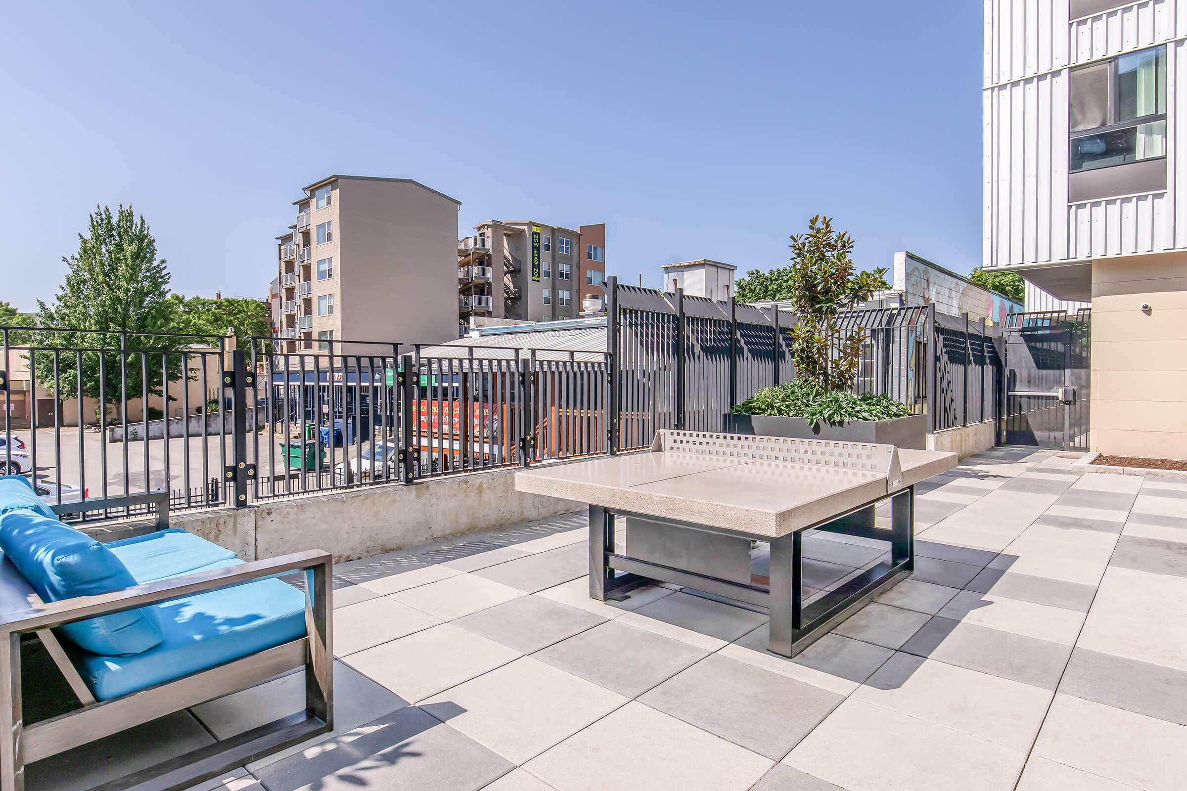 Outdoor seating area featuring a large stone table with benches, surrounded by a fence. The background includes residential buildings and greenery, under a clear blue sky. The space is designed for relaxation and social gatherings.