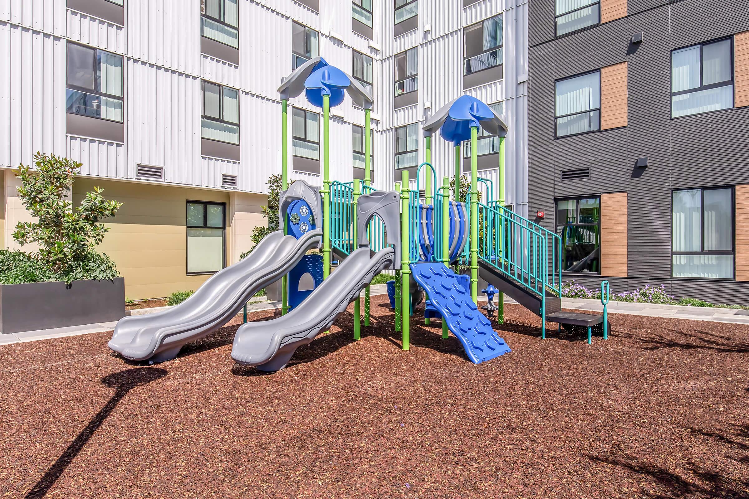 Playground equipment featuring two slides, climbing structures, and various play areas. Surrounded by mulch and located in an outdoor space adjacent to modern buildings with a mix of textures and colors. Ideal for children to play and explore.
