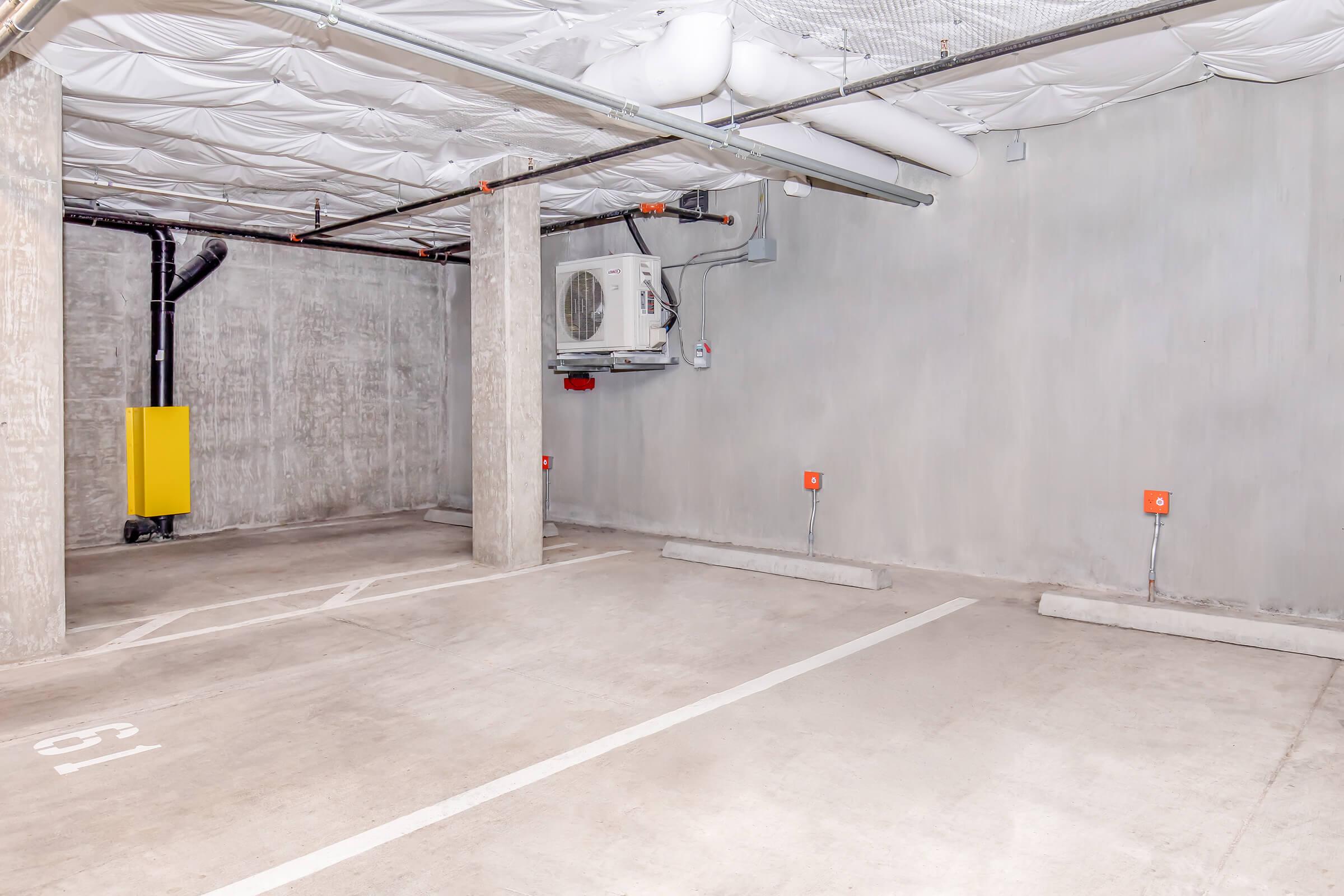 Empty underground parking garage with a concrete floor and walls. There are several marked parking spaces, a yellow emergency water pipe visible, an air conditioning unit mounted on the wall, and minimal lighting. The ceiling features a white, textured surface.
