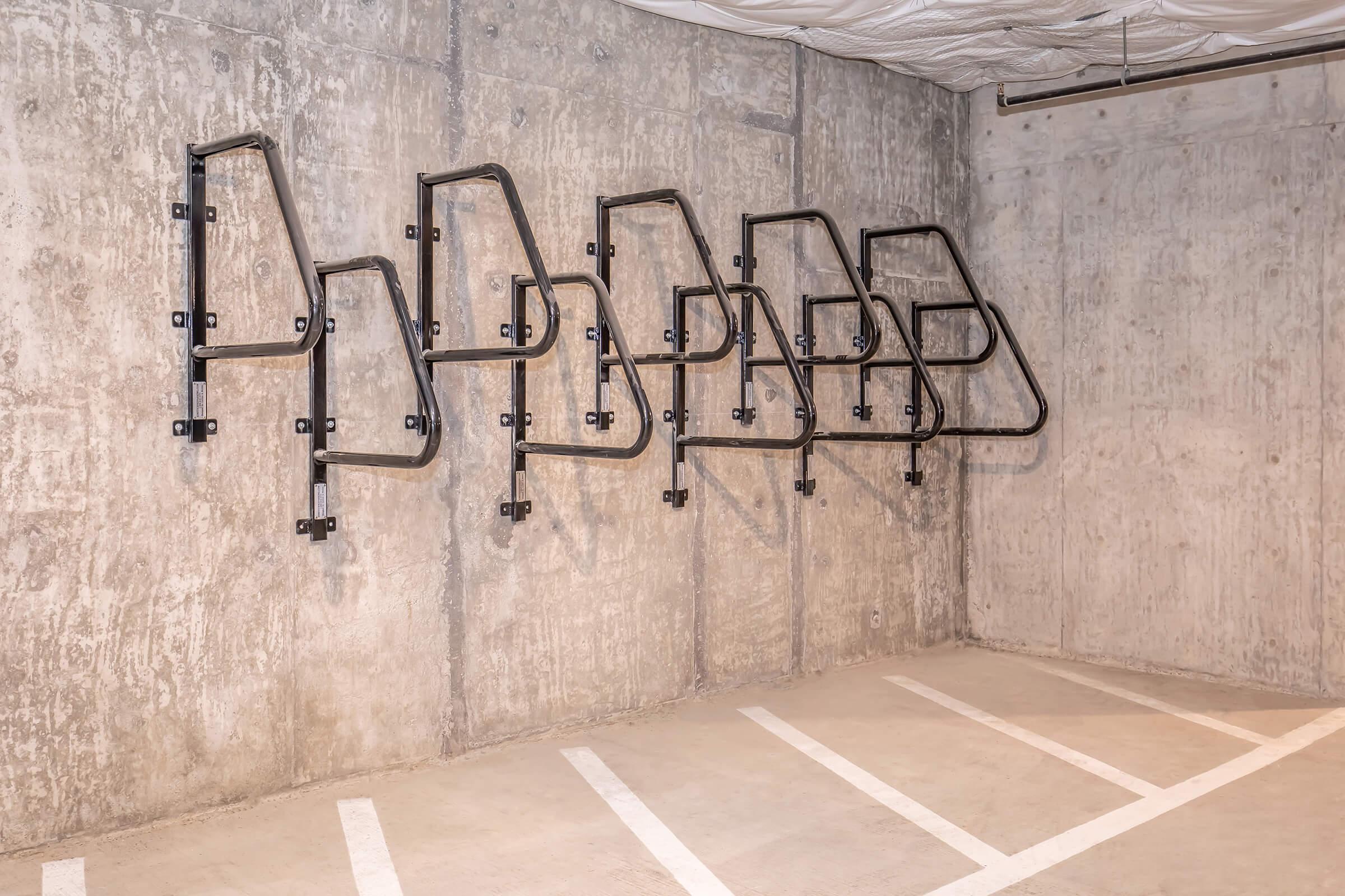 A concrete wall in a parking garage featuring a series of black metal bike racks mounted to the wall, arranged in various positions, with several empty parking spaces visible on the floor. The setting is minimalist and industrial in design.