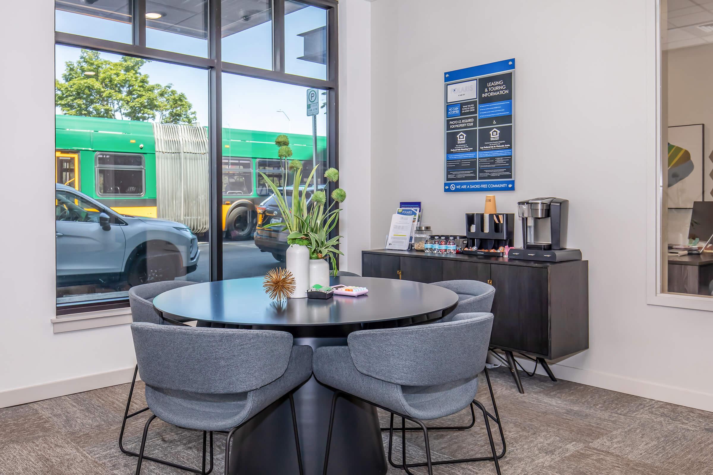 A modern office space featuring a circular black table surrounded by four gray chairs. A small plant centerpiece sits on the table. A coffee station is nearby on a dark cabinet. Large windows offer a view of a street with a green bus. Informational signage is mounted on the wall.