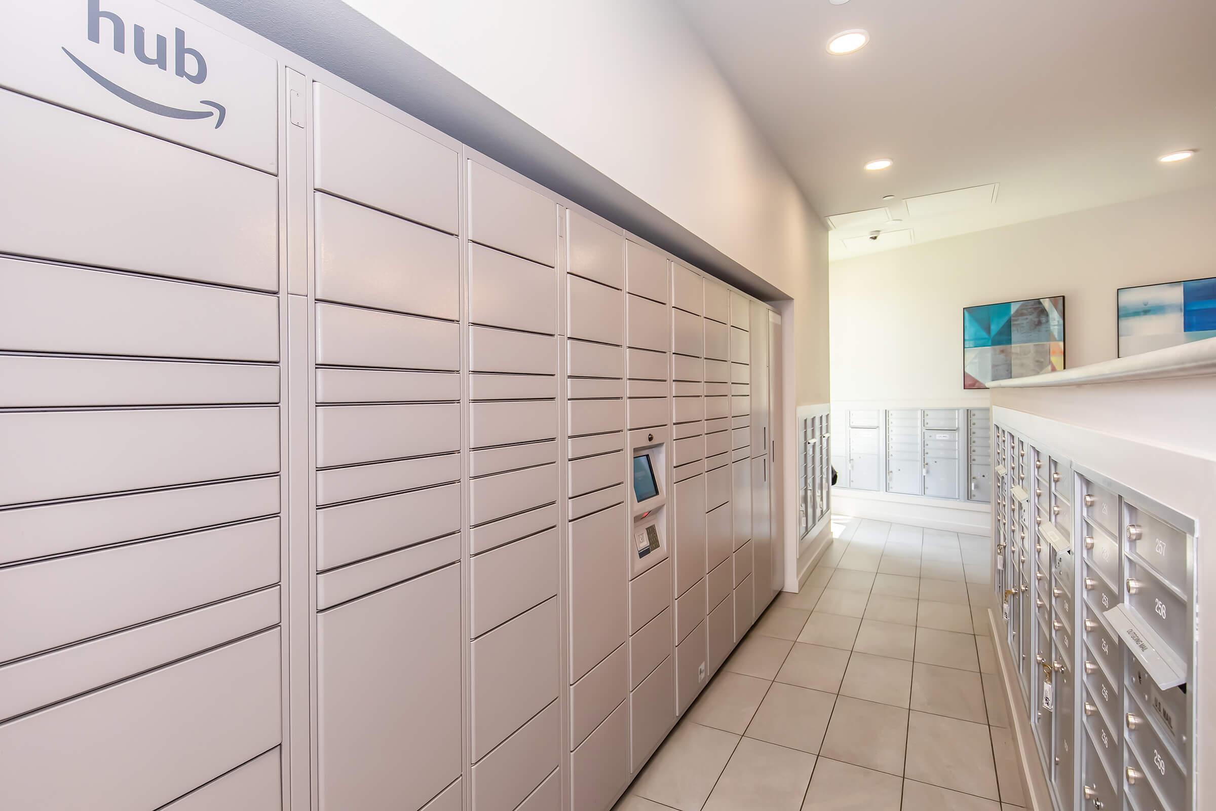 A hallway featuring Amazon package lockers alongside traditional mailboxes. The space is well-lit with modern design elements, including artwork on the walls. The lockers are arranged in rows, allowing for secure package delivery and retrieval.
