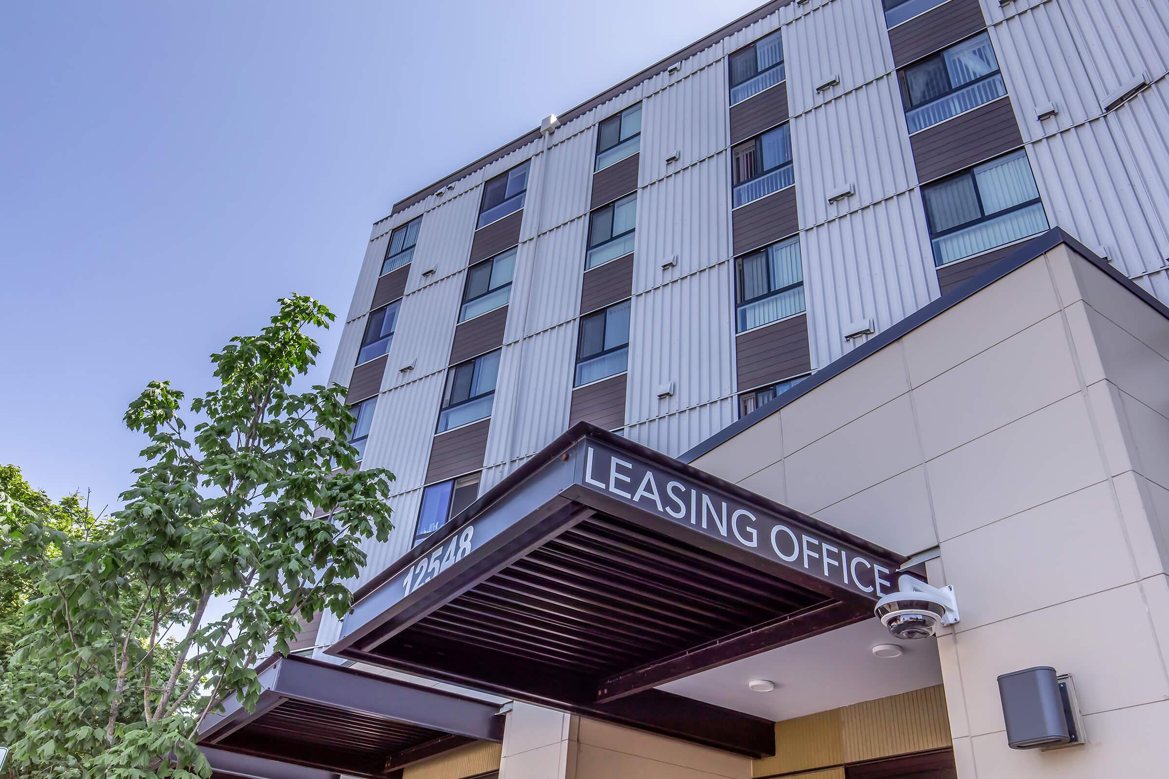 A modern leasing office building with a prominent sign reading "LEASING OFFICE." The structure features a mix of materials, large windows, and a clean entrance. A tree is visible in the foreground, adding greenery to the urban setting under a clear blue sky.