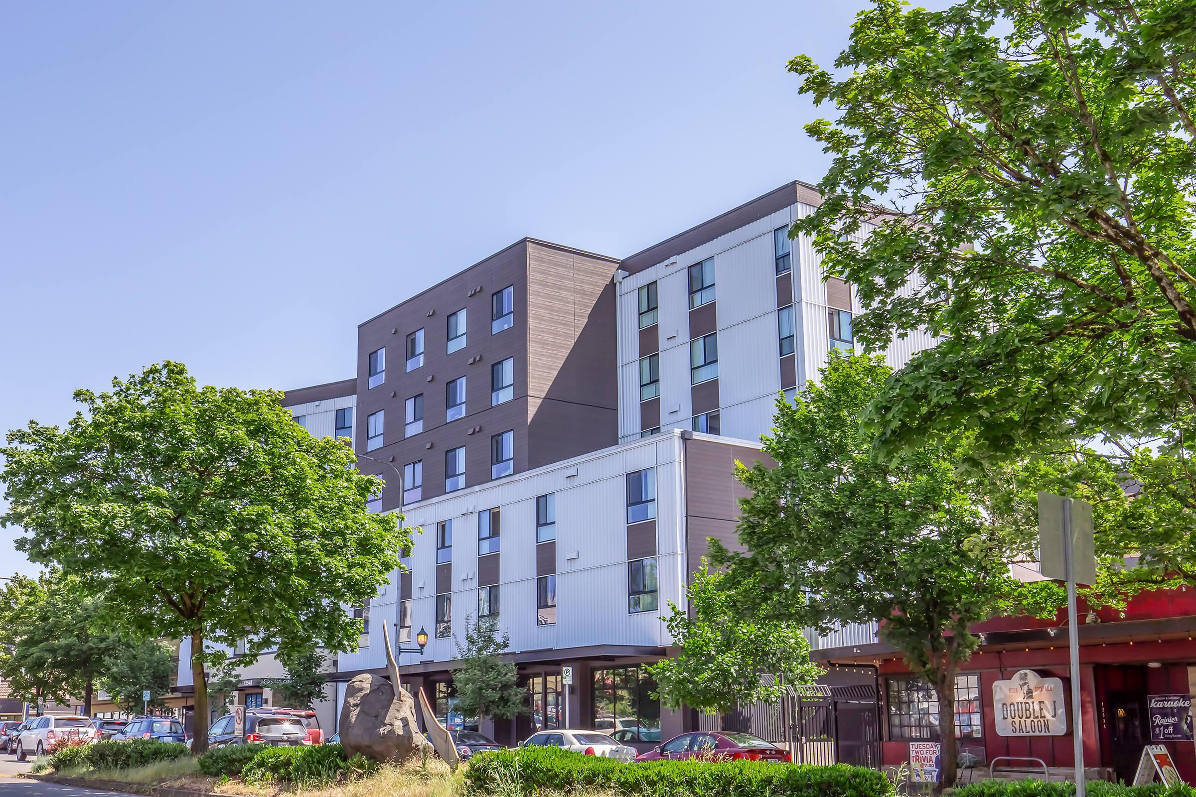 A modern multi-story building with a mix of dark and light-colored panels. The structure features large windows, and there are several trees and greenery in the foreground. In front, there is a small restaurant or shop with a sign that indicates its name. The sky is clear and blue.