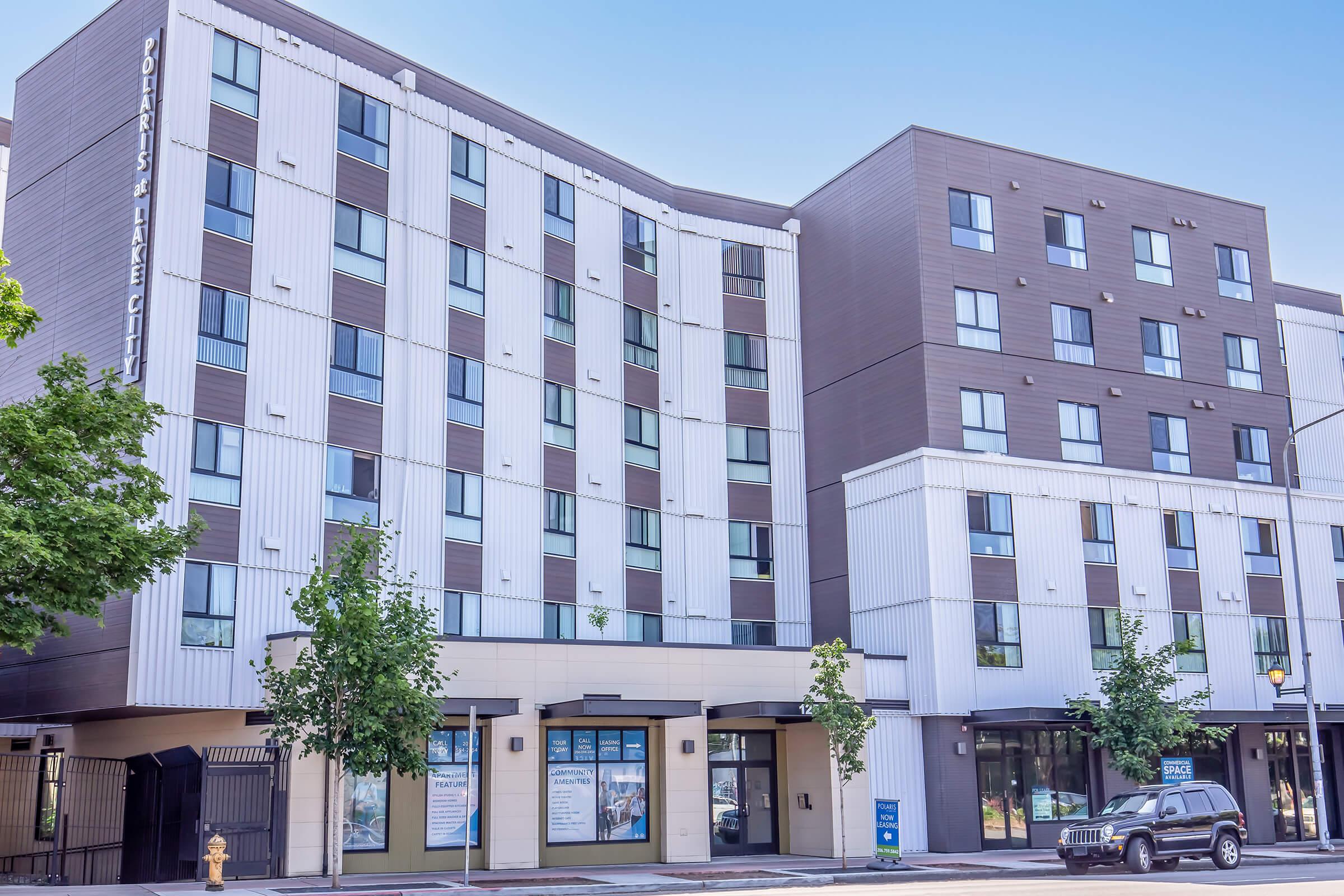 A modern hotel building with a mix of light and dark exterior panels, featuring multiple windows and trees in front. The ground floor has large windows displaying signage for the hotel, with a black vehicle parked nearby. The sky is clear and blue, creating a bright atmosphere.