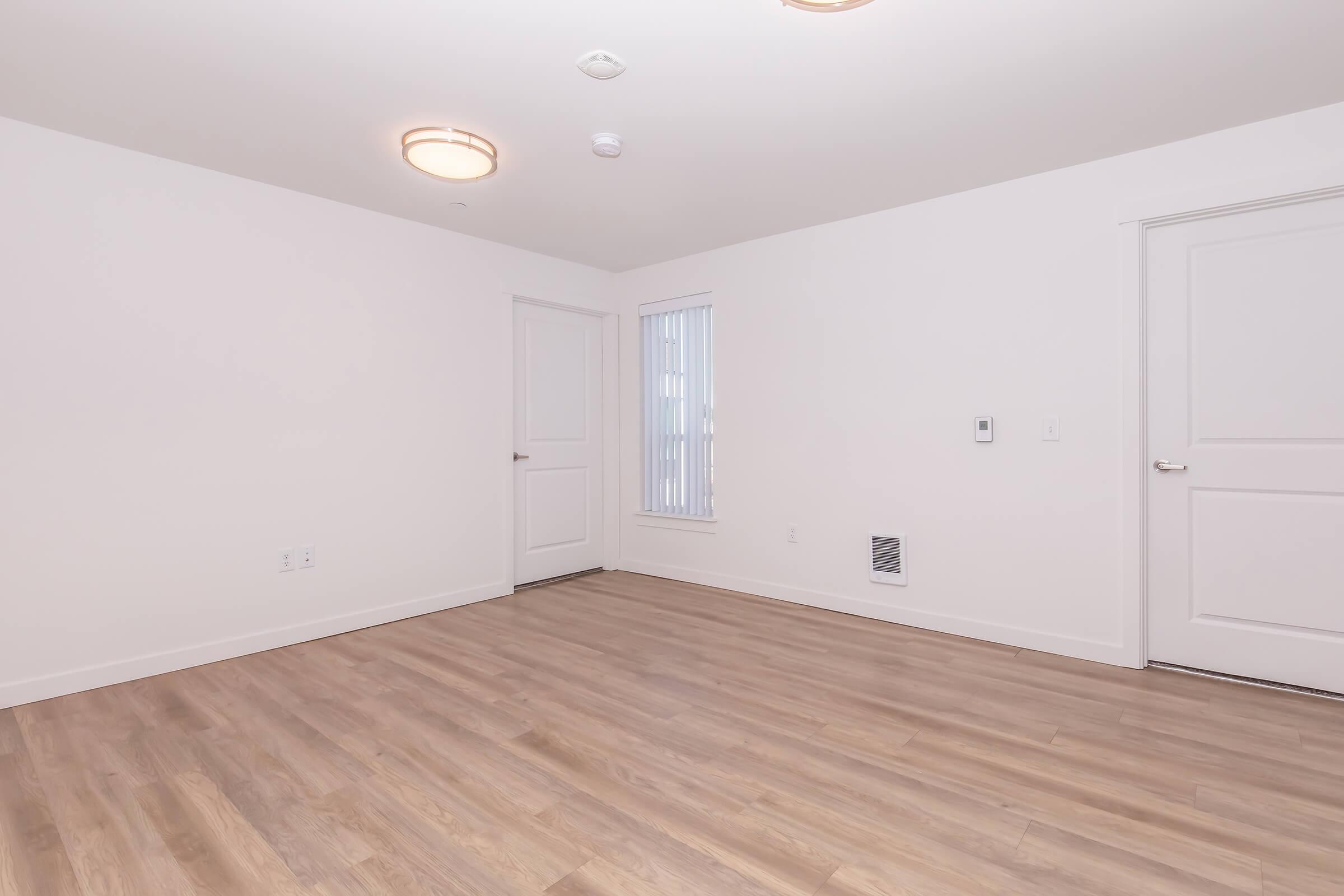 Empty room with light-colored walls and hardwood flooring. Contains a single window with vertical blinds, a door on one side, and a vent on the floor. The ceiling features recessed lighting and two modern light fixtures. The overall space appears clean and well-lit, suitable for various furnishings.