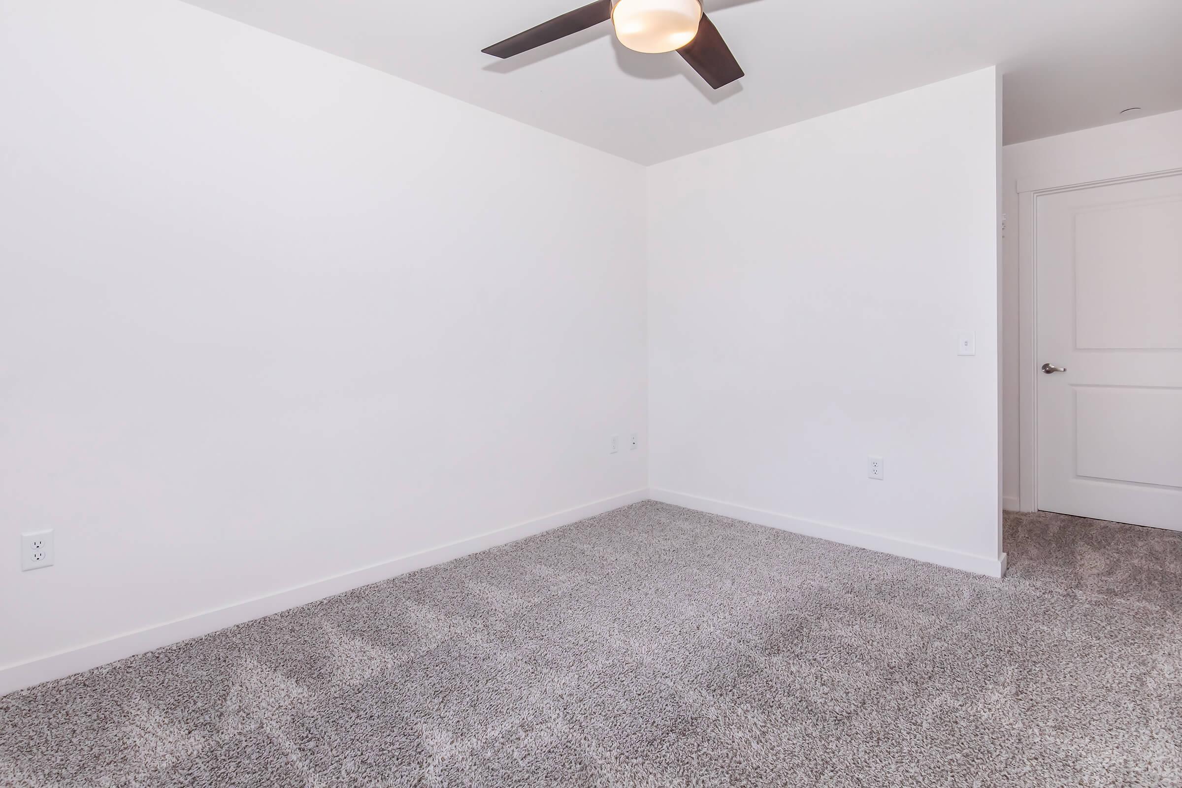 A spacious empty room with light-colored walls, a ceiling fan, and beige carpet. The room features a single door leading to another area, providing a clean and uncluttered look, perfect for personalization or decoration. Natural light may enter through the door, enhancing the bright atmosphere.