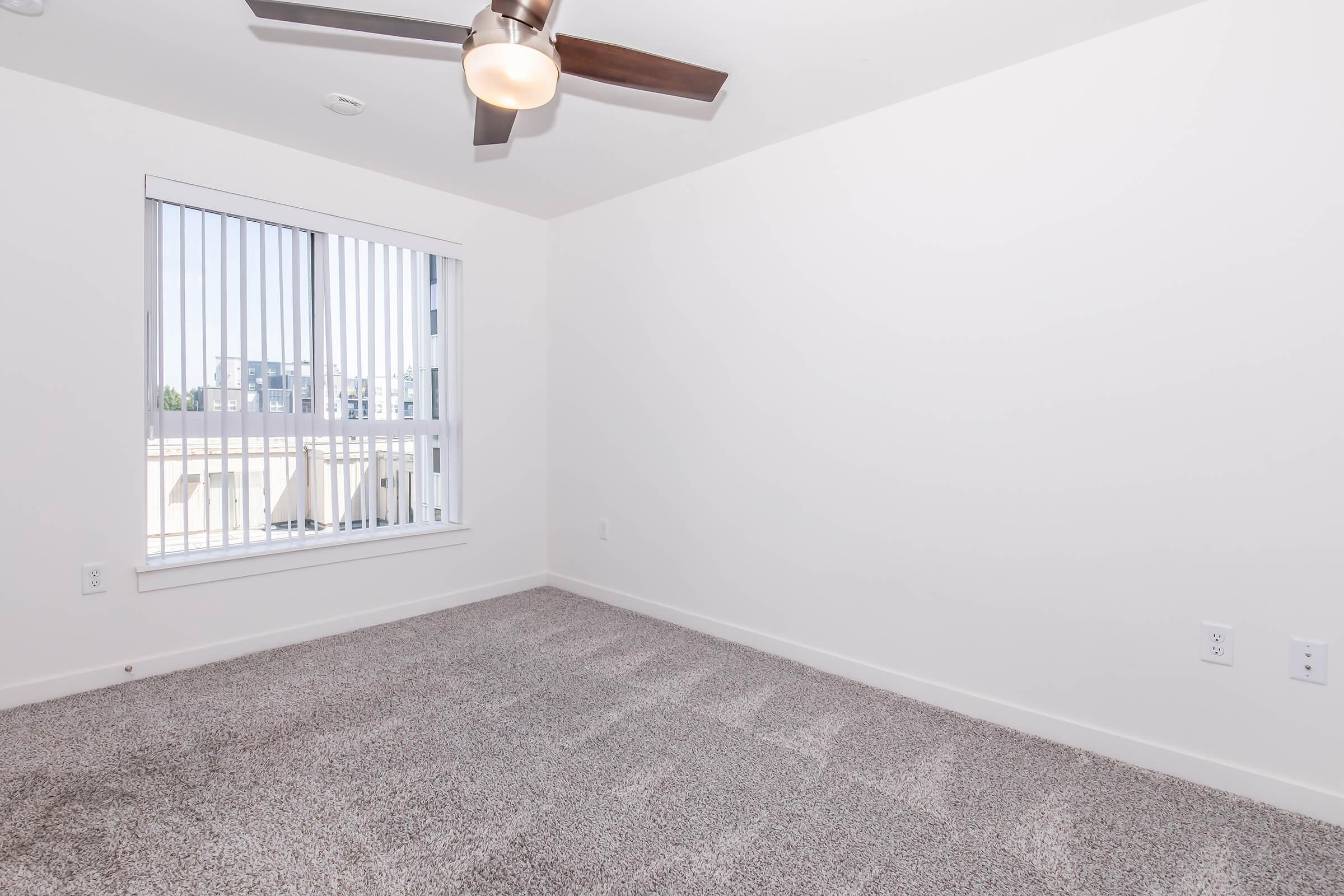A spacious, empty room with light gray carpet and white walls. It features a modern ceiling fan and a large window with vertical blinds allowing natural light to enter. The room is unadorned, giving a clean and minimalist appearance, ideal for various decorating possibilities.