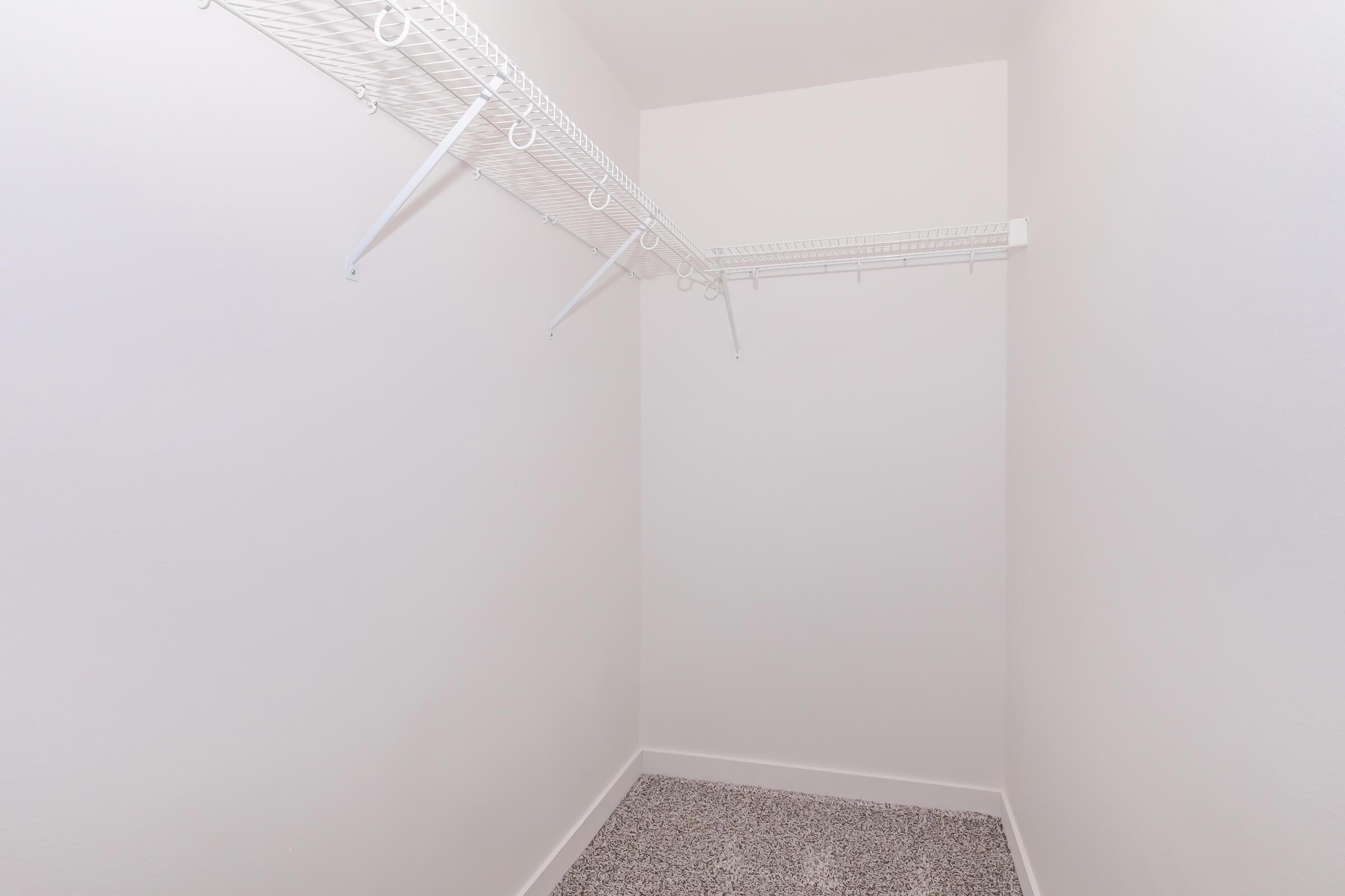 A small, empty closet with white walls and a light gray carpet. There are two wire shelving units mounted on the wall, without any items stored. The space is well-lit and devoid of personal belongings.