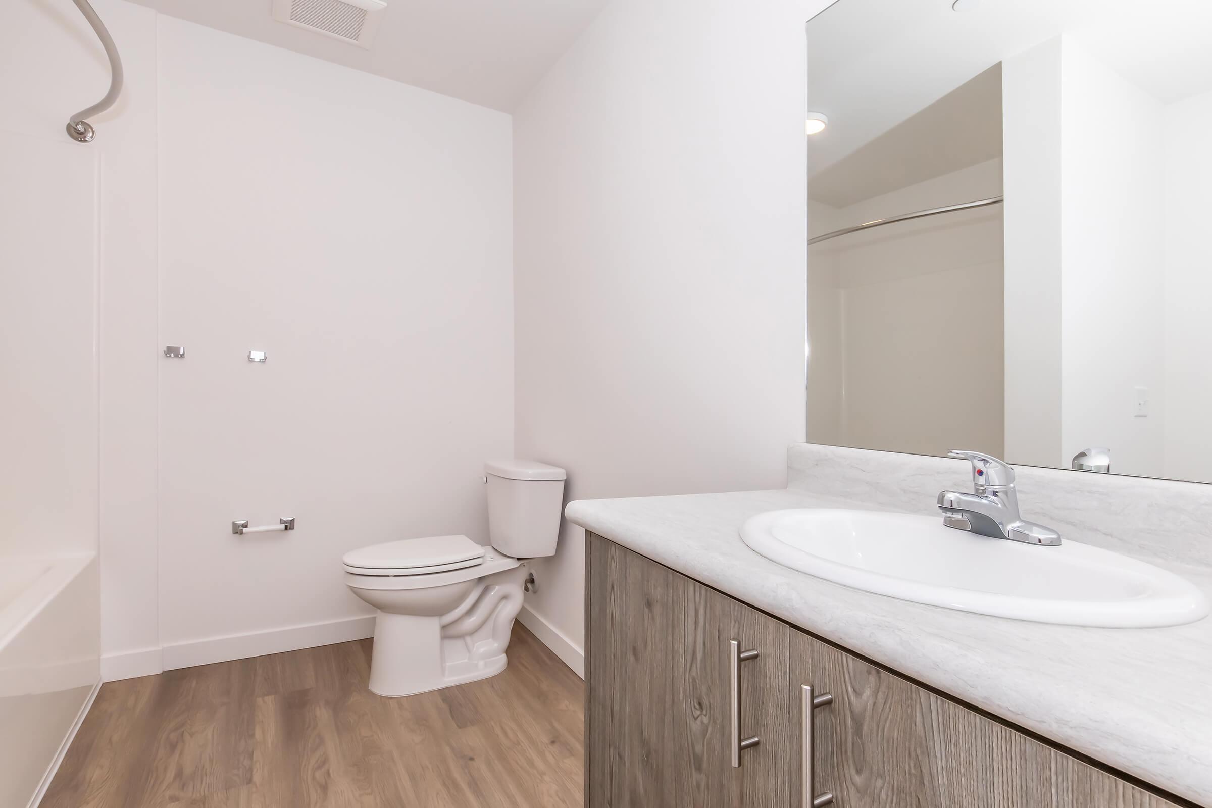 A modern bathroom featuring a clean design. It includes a bathtub on the left, a toilet, and a vanity with a sink. The walls are painted white, and the floor is made of light wood. A mirror above the sink reflects the space, adding to the brightness of the room.