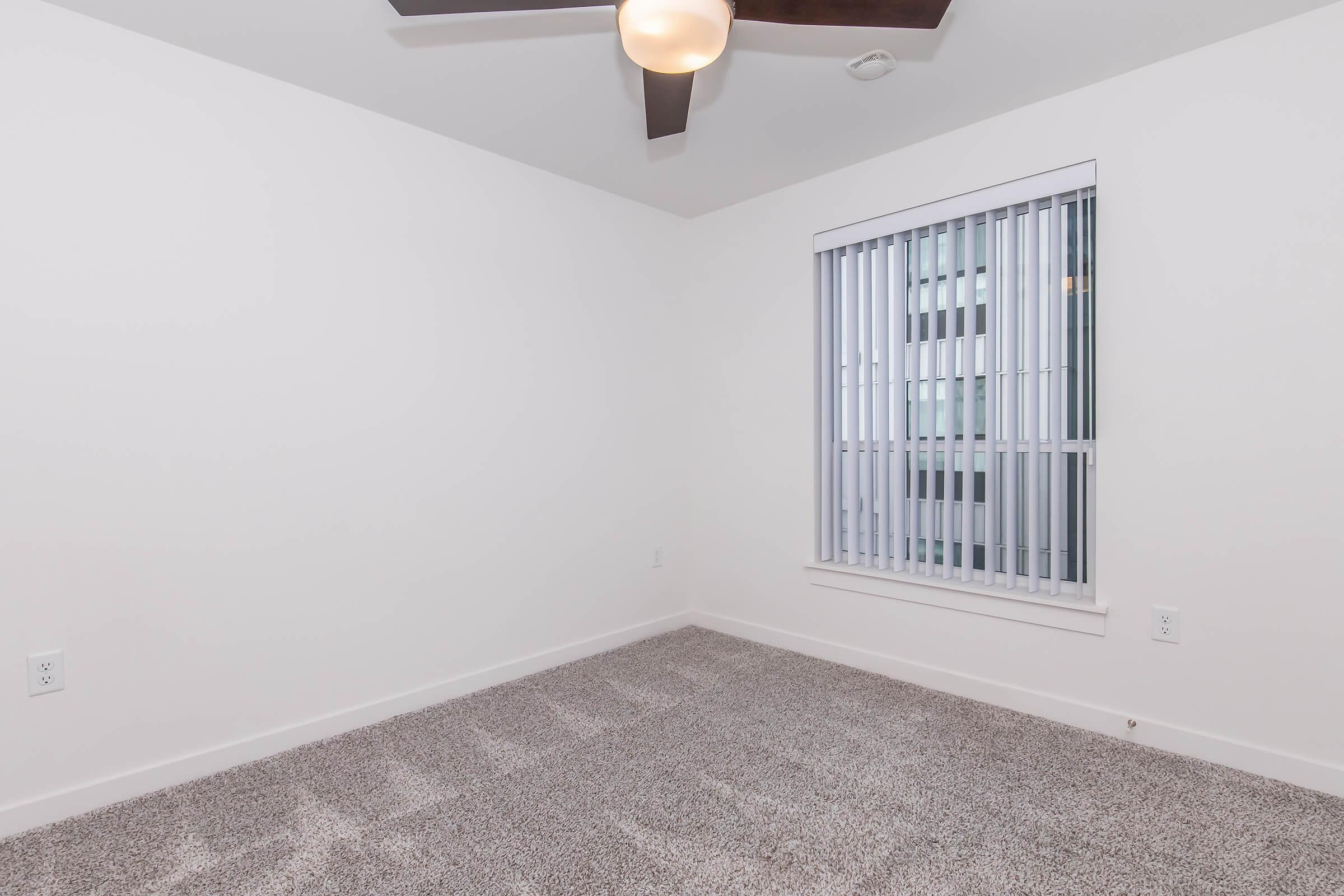 A sparsely furnished room featuring light-colored walls and a ceiling fan. The floor is covered with beige carpet, and a window with vertical blinds is visible, allowing some natural light. The overall atmosphere is bright and simple, suggesting a clean and modern living space.