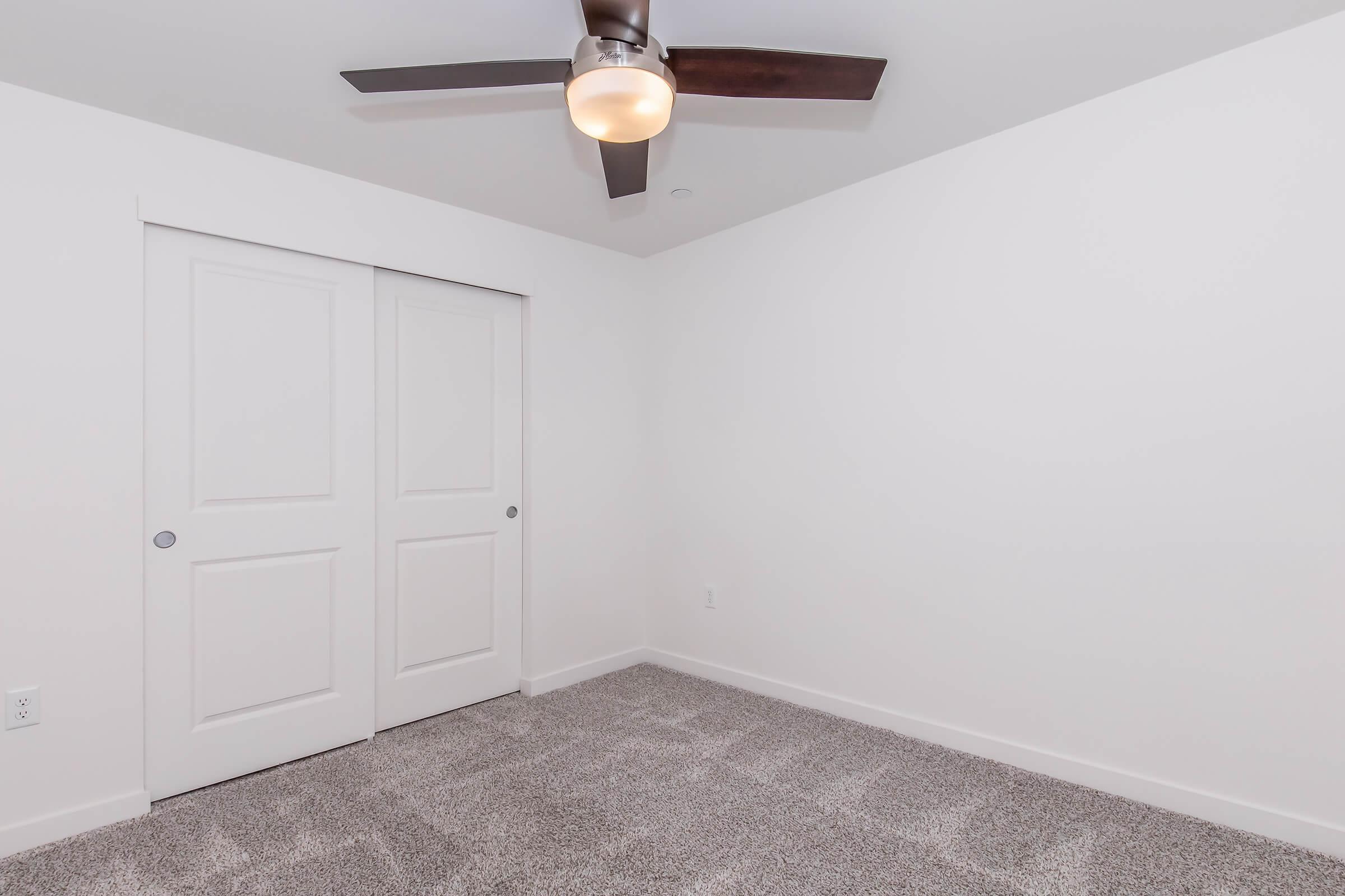 Empty room featuring light gray carpet and white walls. A ceiling fan with a light fixture is installed in the center, and there are double sliding doors on one side, leading to a closet or storage space. The room is well-lit and has a clean, modern aesthetic.