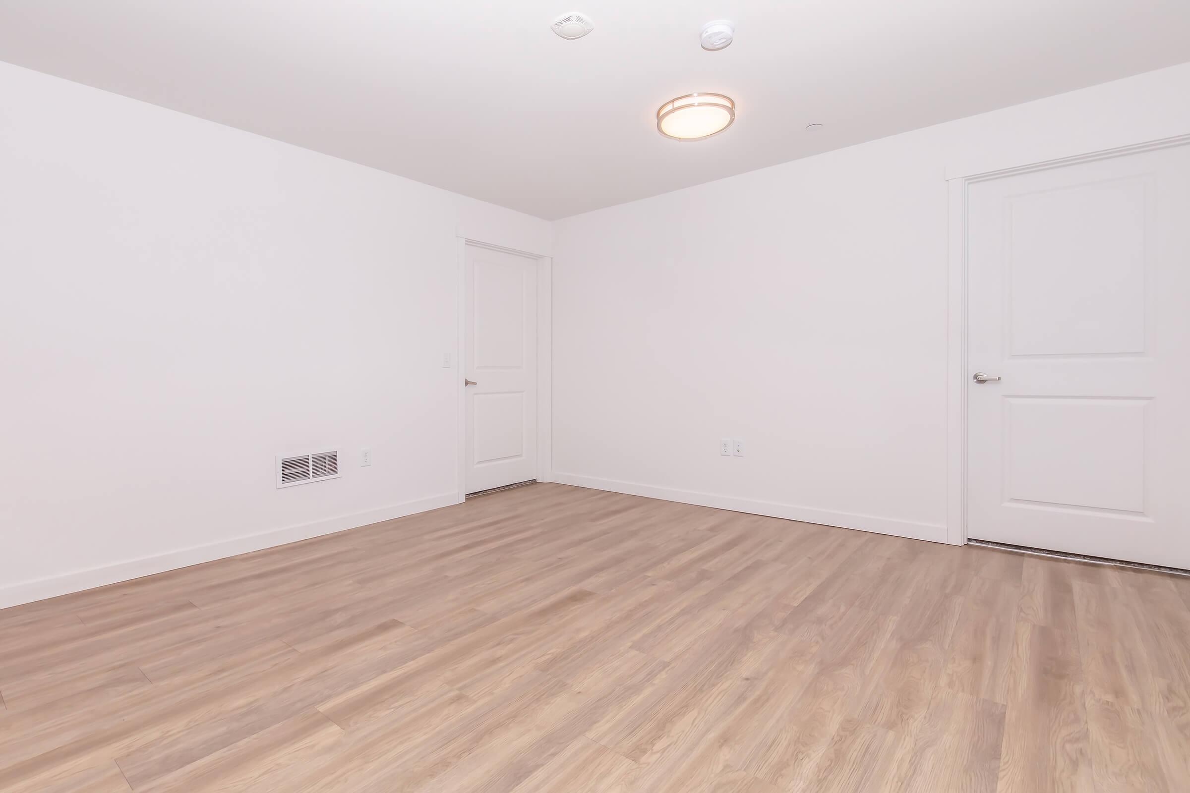 Empty room with white walls and a natural wood floor. A single ceiling light fixture is visible. There are two doors, one on the right and one on the left, leading to adjacent spaces. The room has a clean, minimalist aesthetic with ample natural light.