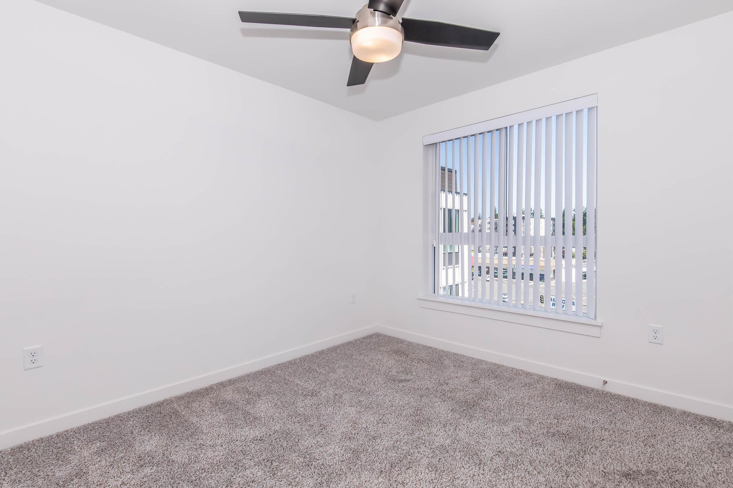 A vacant room with light gray carpet, a ceiling fan, and white walls. There is a large window with vertical blinds allowing natural light to enter, revealing a view outside. The space is unfurnished and appears bright and airy.