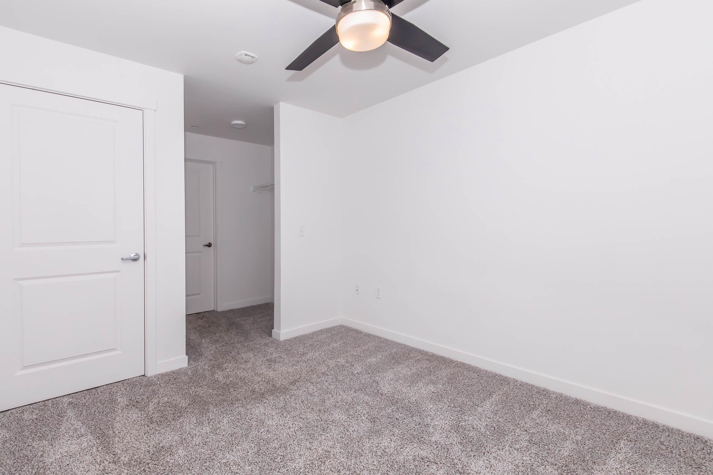 An empty room with light gray carpet and white walls. A ceiling fan is mounted on the ceiling, and there is a closed white door on the left and a doorway leading to another area on the right. The space is well-lit and minimalistic, suitable for various uses.