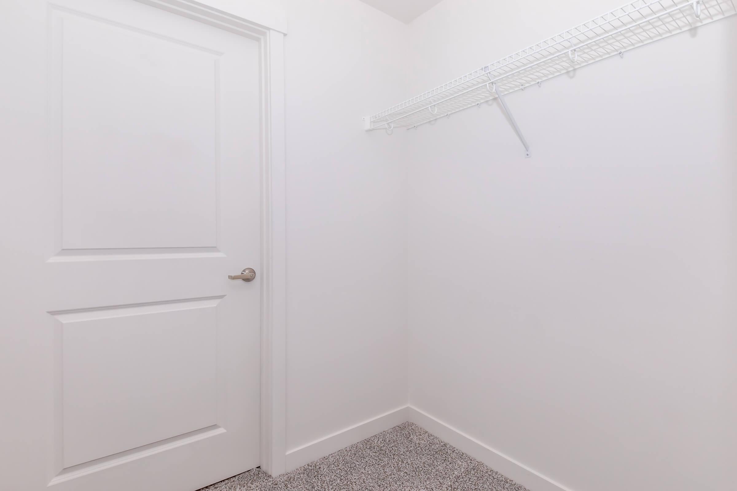 Empty room with plain white walls and a light gray carpet. There is a white door on the left and a wire shelving unit mounted on the right wall, creating a minimalistic and spacious appearance. The overall ambiance is clean and bright, suggesting a closet or storage area.