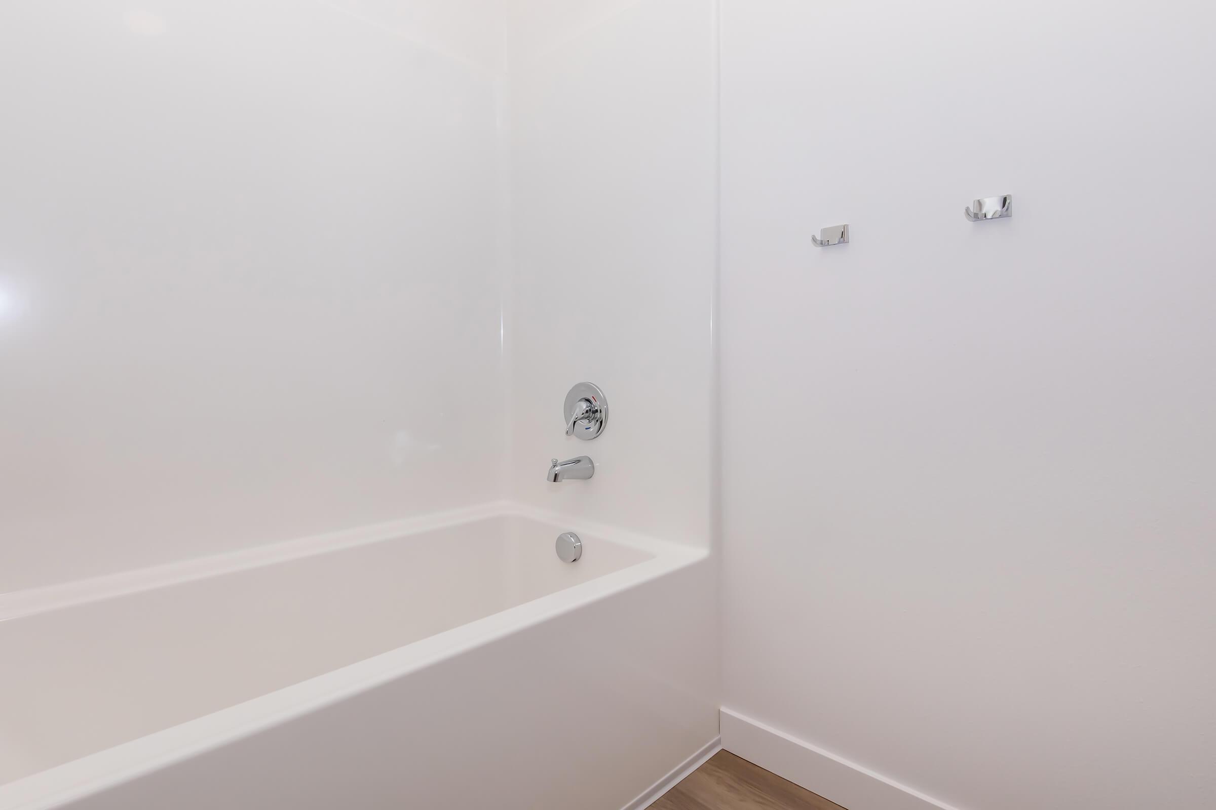 A clean, modern bathtub in a minimalistic bathroom setting. The tub is white and built into a white wall with a single faucet and showerhead. Two empty towel hooks are mounted on the wall beside the tub, and the floor features light-colored wood. Natural light reflects softly on the surfaces.