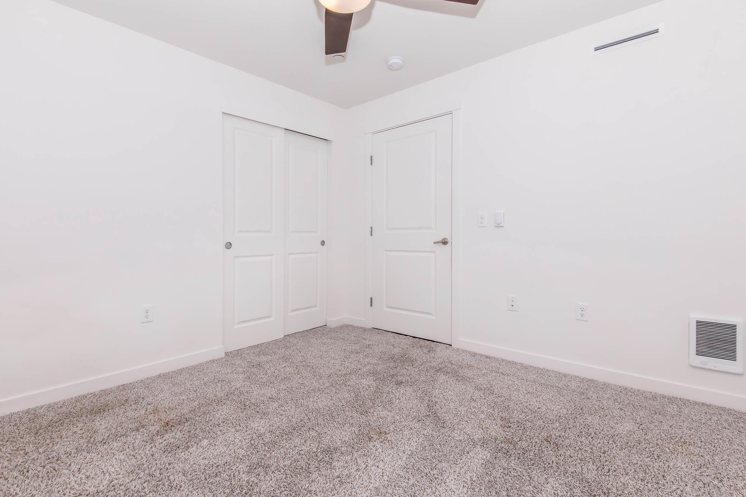 Empty room with light-colored walls and a ceiling fan. The floor is covered in light gray carpet. One wall features a closed door with a simple panel design, while another wall has a door leading to a closet. The overall space is bright and minimalistic.