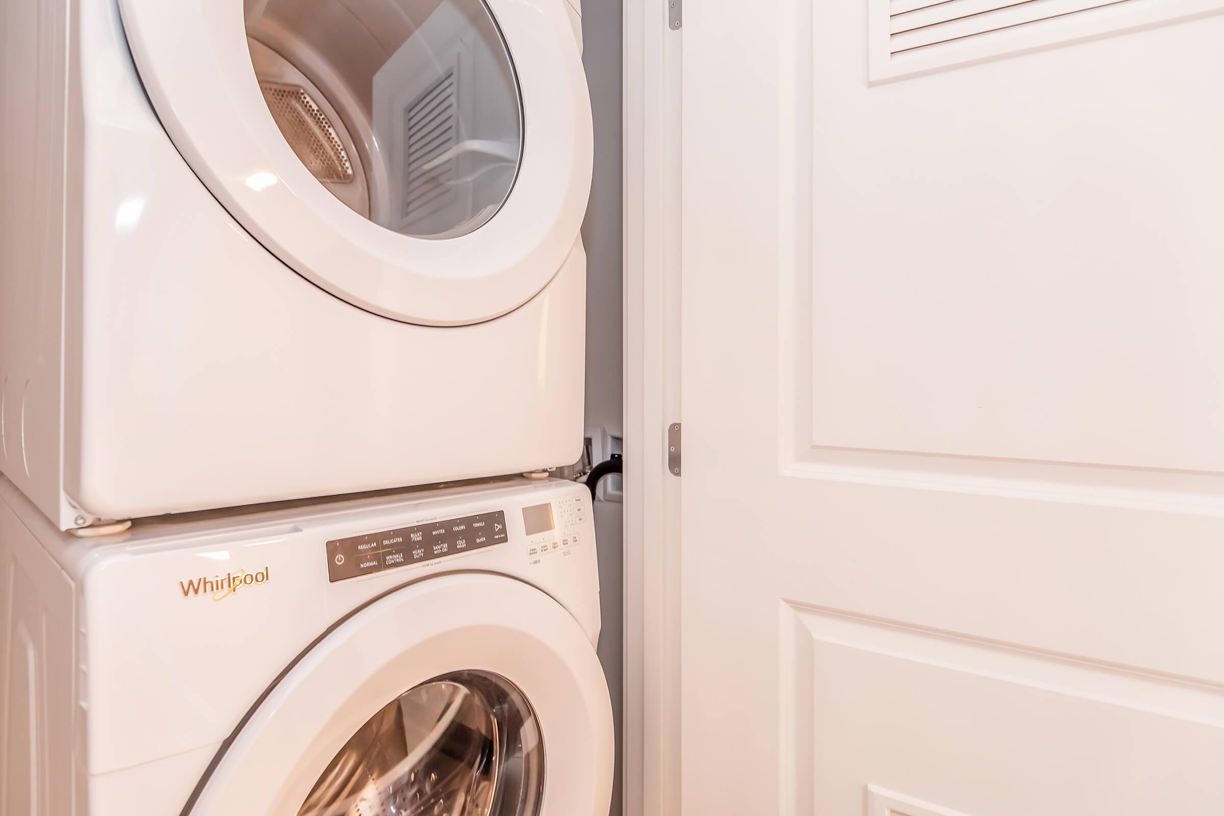 A stackable Whirlpool washer and dryer are positioned next to a white door in a laundry area. The appliances have large circular doors, and the control panel on the front of the washer features various buttons and settings. The space is designed for efficient use, with the door providing access to the area.