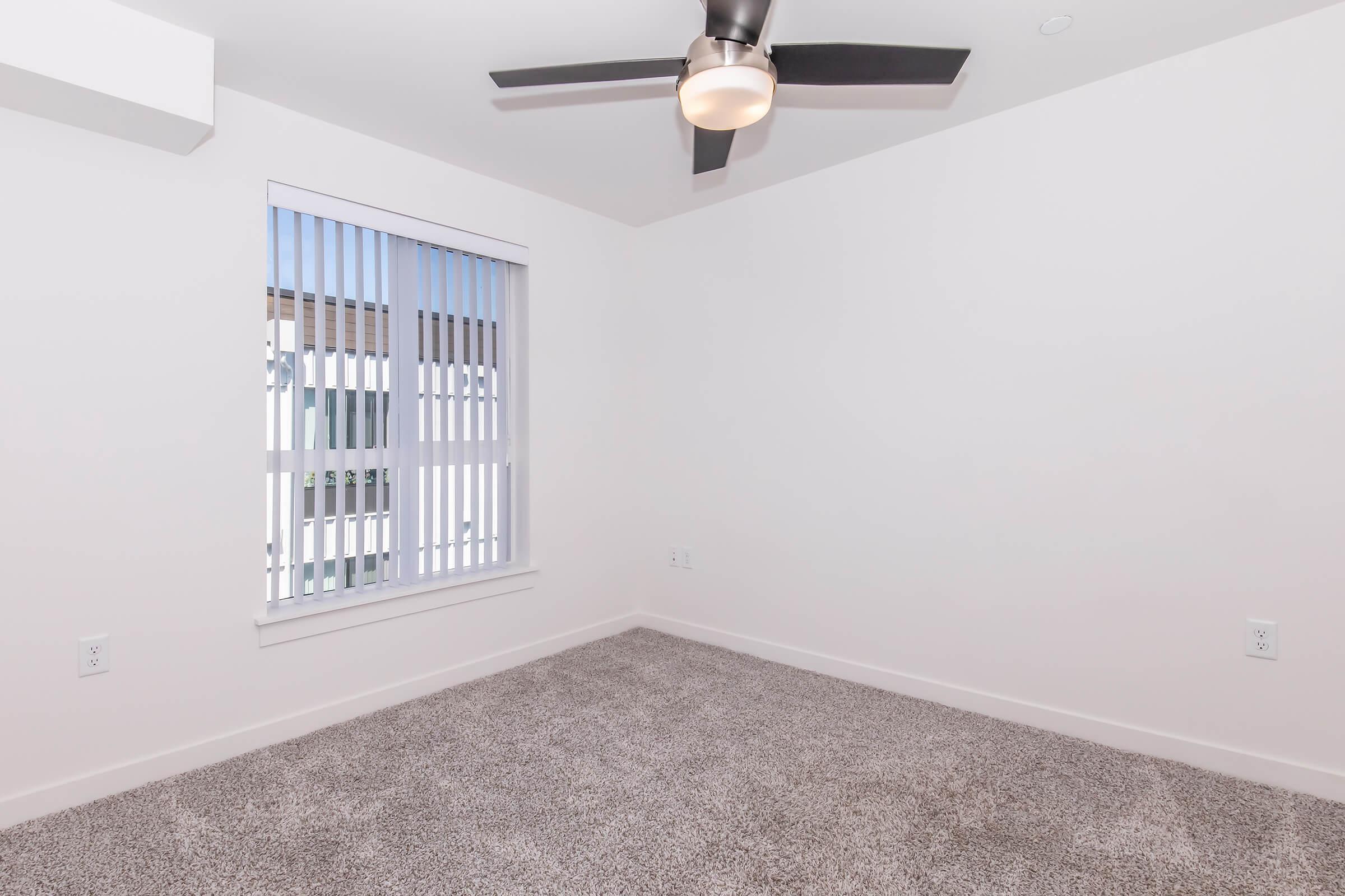 A well-lit room with light gray carpet flooring and white walls. It features a ceiling fan and a window with vertical blinds, allowing natural light to enter. The room is empty, creating a spacious and clean appearance.