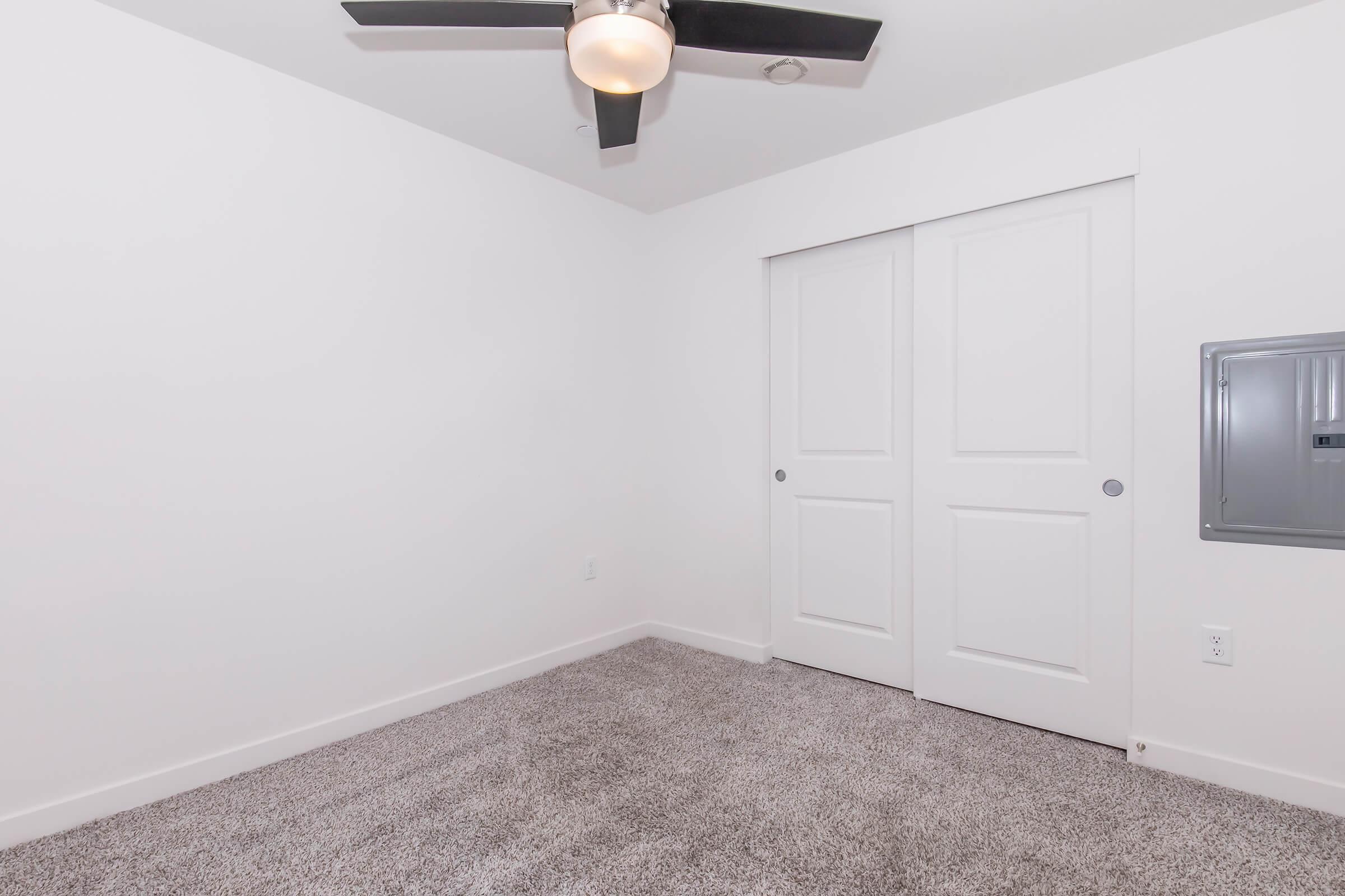 A bright, empty room with light gray carpet and plain white walls. A ceiling fan is mounted on the ceiling, and there are two closed closet doors on one side. A small mailbox is visible near the floor on another wall. The space is uncluttered, offering a minimalist aesthetic.