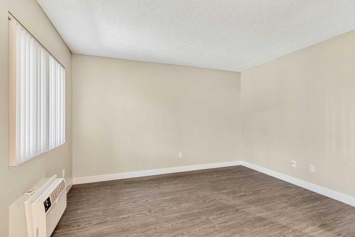 Empty room with beige walls and a light-colored ceiling. The floor is covered in wood-like laminate. There is a window with vertical blinds on one side, and an air conditioning unit mounted on the wall. The room is well-lit, creating a spacious and clean appearance.