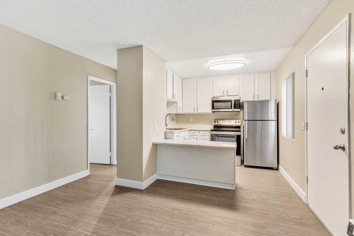 Bright and modern kitchen featuring white cabinetry, stainless steel appliances, and a spacious countertop. The open layout connects to a living area with neutral-colored walls and wood-like flooring. A doorway leads to another room, and a front door is visible on the right. Natural light enters through a window.