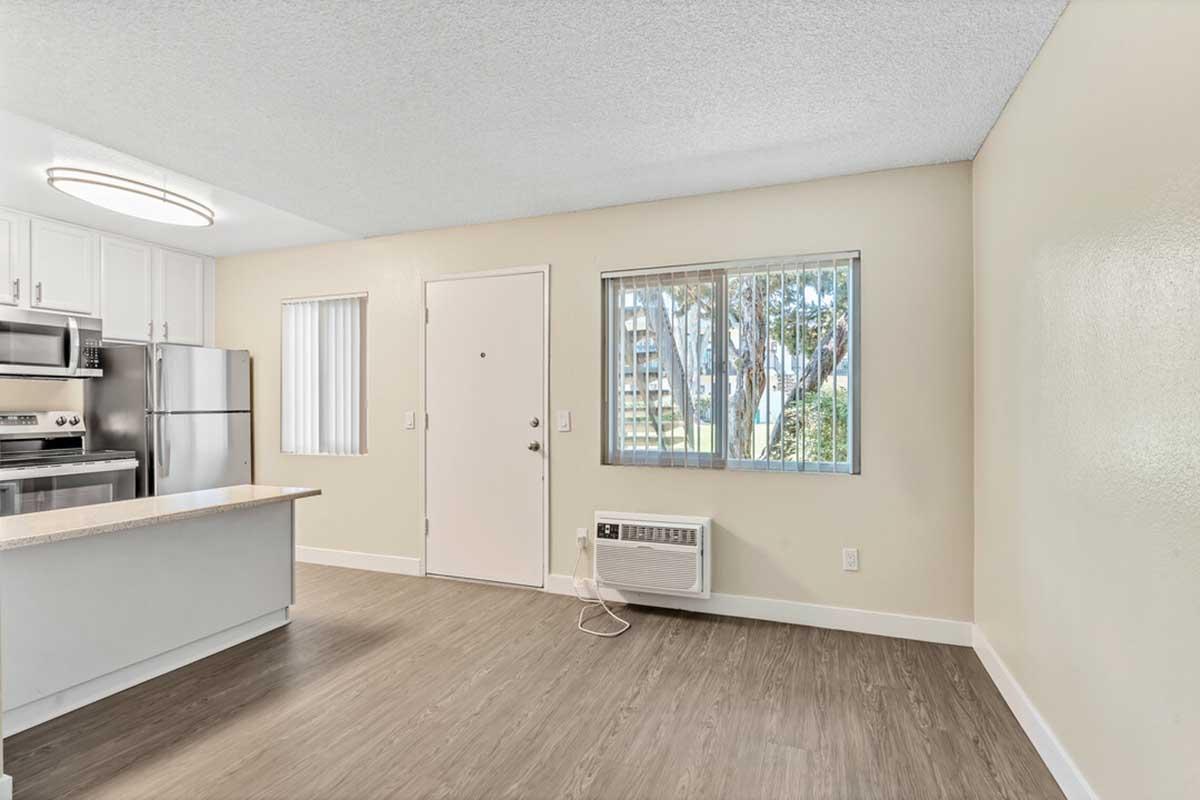An interior view of a modern, bright kitchen and living area featuring stainless steel appliances, a window with blinds, a door, and an air conditioning unit. The flooring is a light wood laminate, and the walls are painted in a soft neutral color.