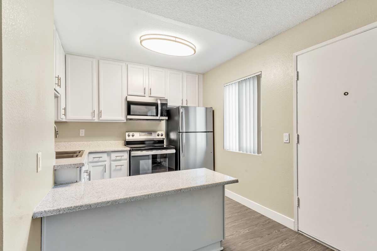 Modern kitchen featuring white cabinetry, stainless steel appliances, including a microwave and refrigerator, and a countertop with bar-style seating. The walls are a light shade, and there is a window with vertical blinds. A round ceiling light illuminates the space, creating a bright atmosphere.