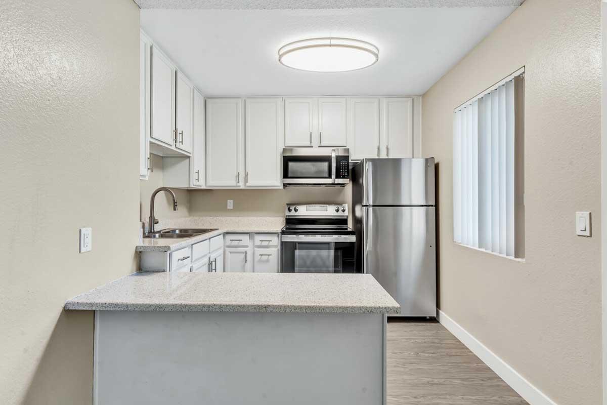 Modern kitchen featuring white cabinetry, stainless steel appliances including an oven and refrigerator, a quartz countertop, and a pendant light fixture overhead. The space is well-lit with natural light streaming in through a window with vertical blinds. The flooring is a light wood laminate.